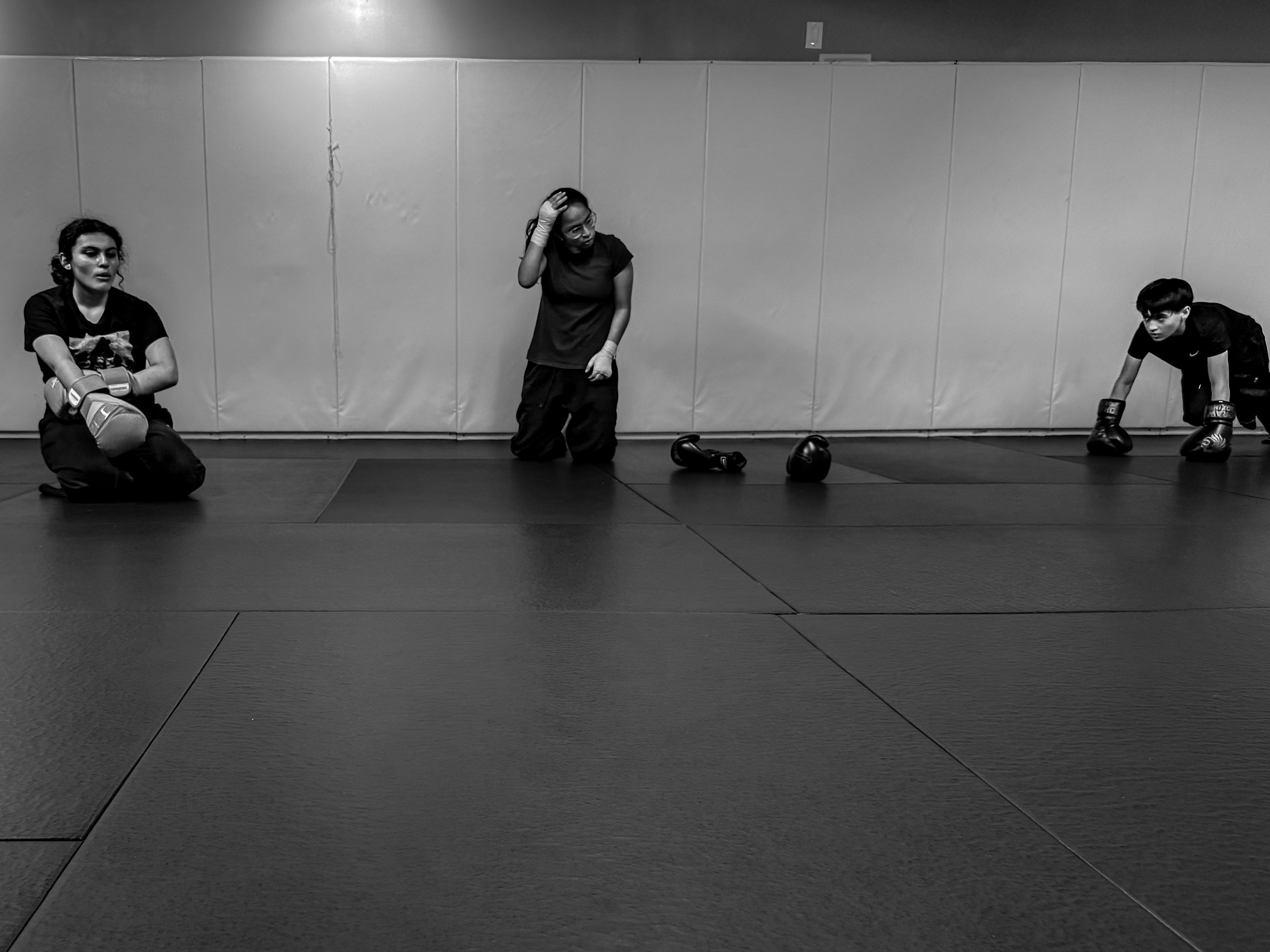 Three women in a boxing gym, one sitting on the floor with boxing gloves, one kneeling adjusting gloves, and one crouching ready to box.