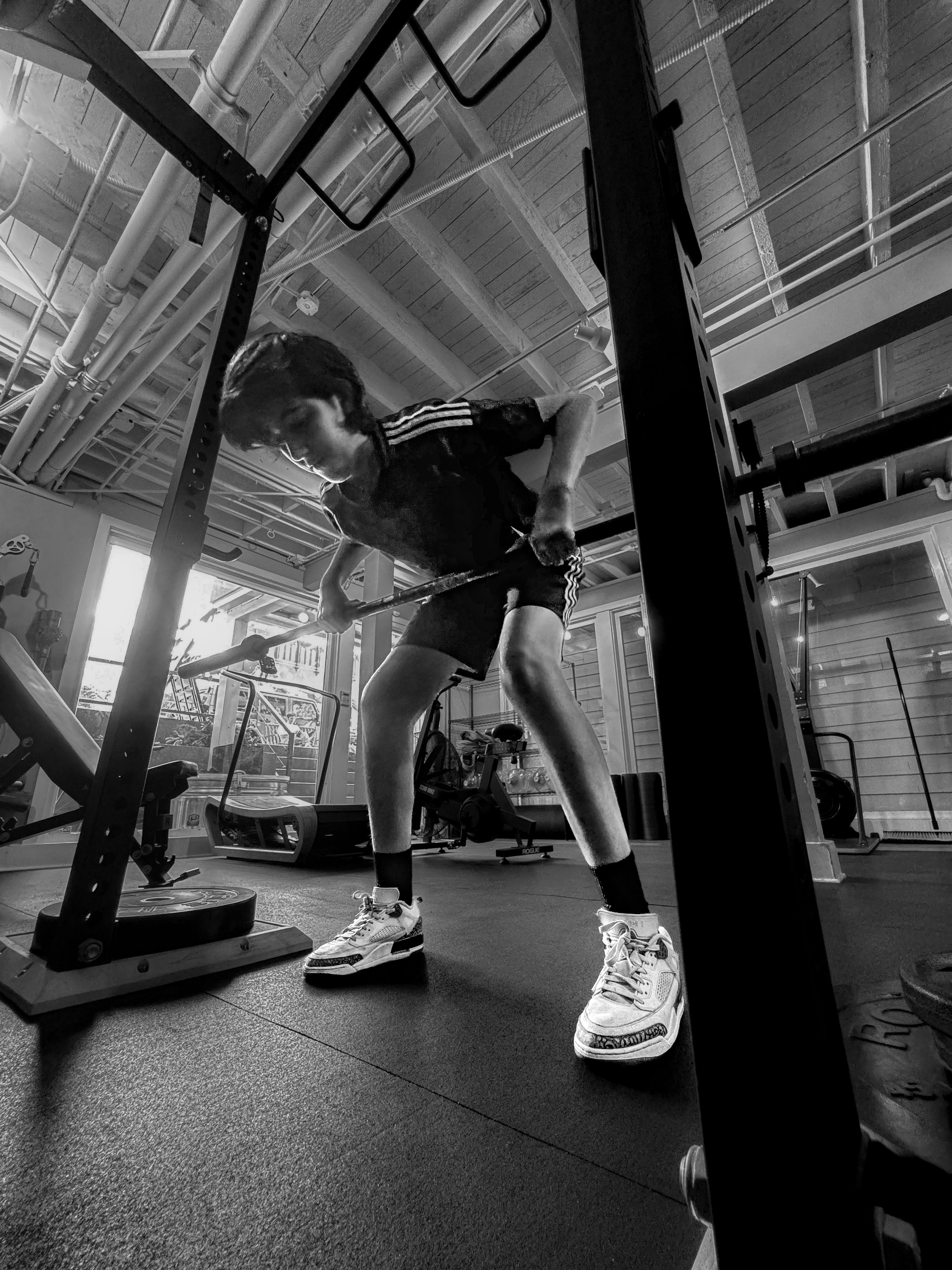 A man in athletic clothing lifts a barbell with weights in a gym with high ceilings, exposed pipes, and large windows in the background.