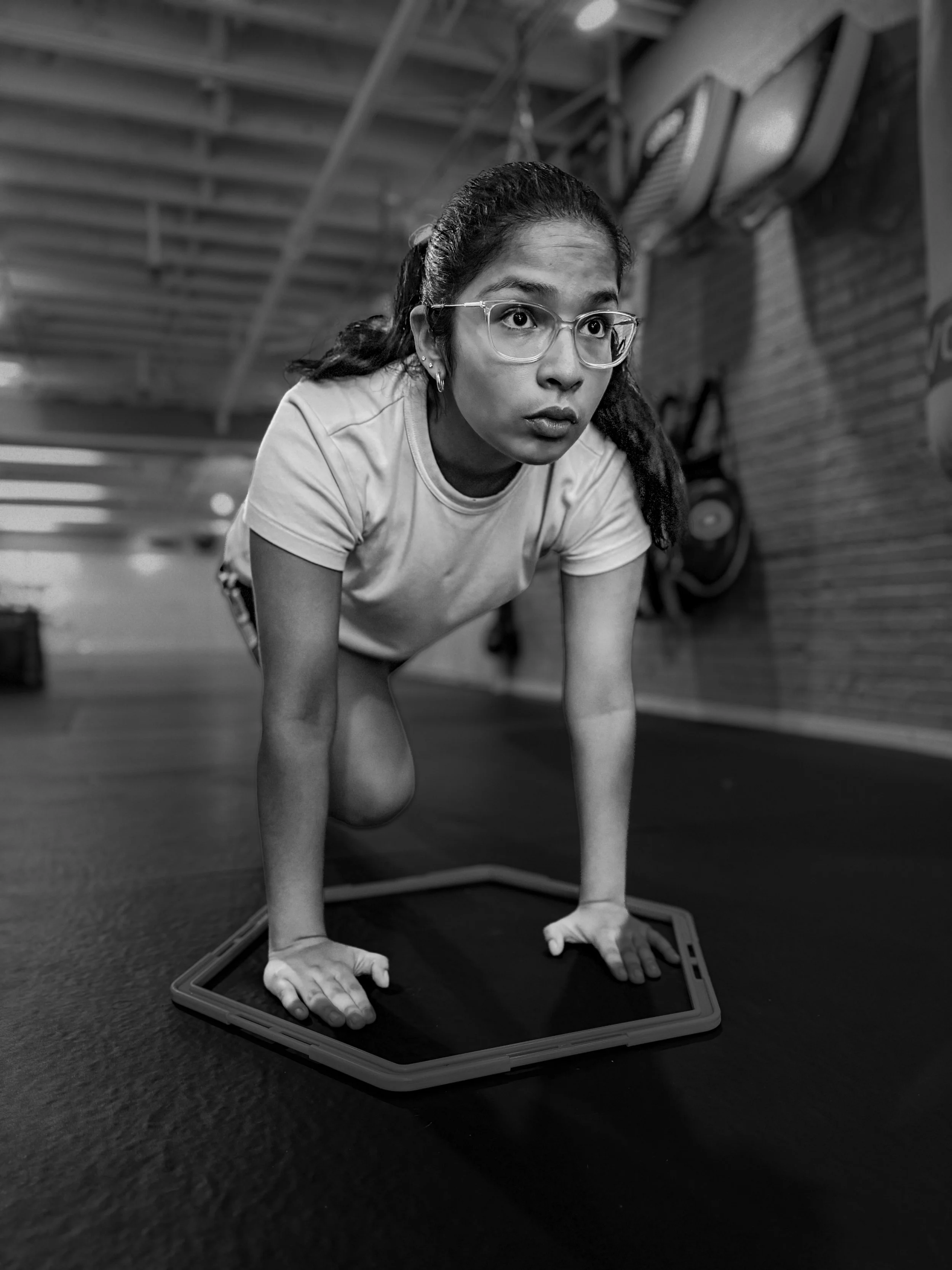 A woman with glasses and long dark hair in a ponytail doing a yoga pose on a mat, with her hands and feet on the mat, in a gym or studio setting.