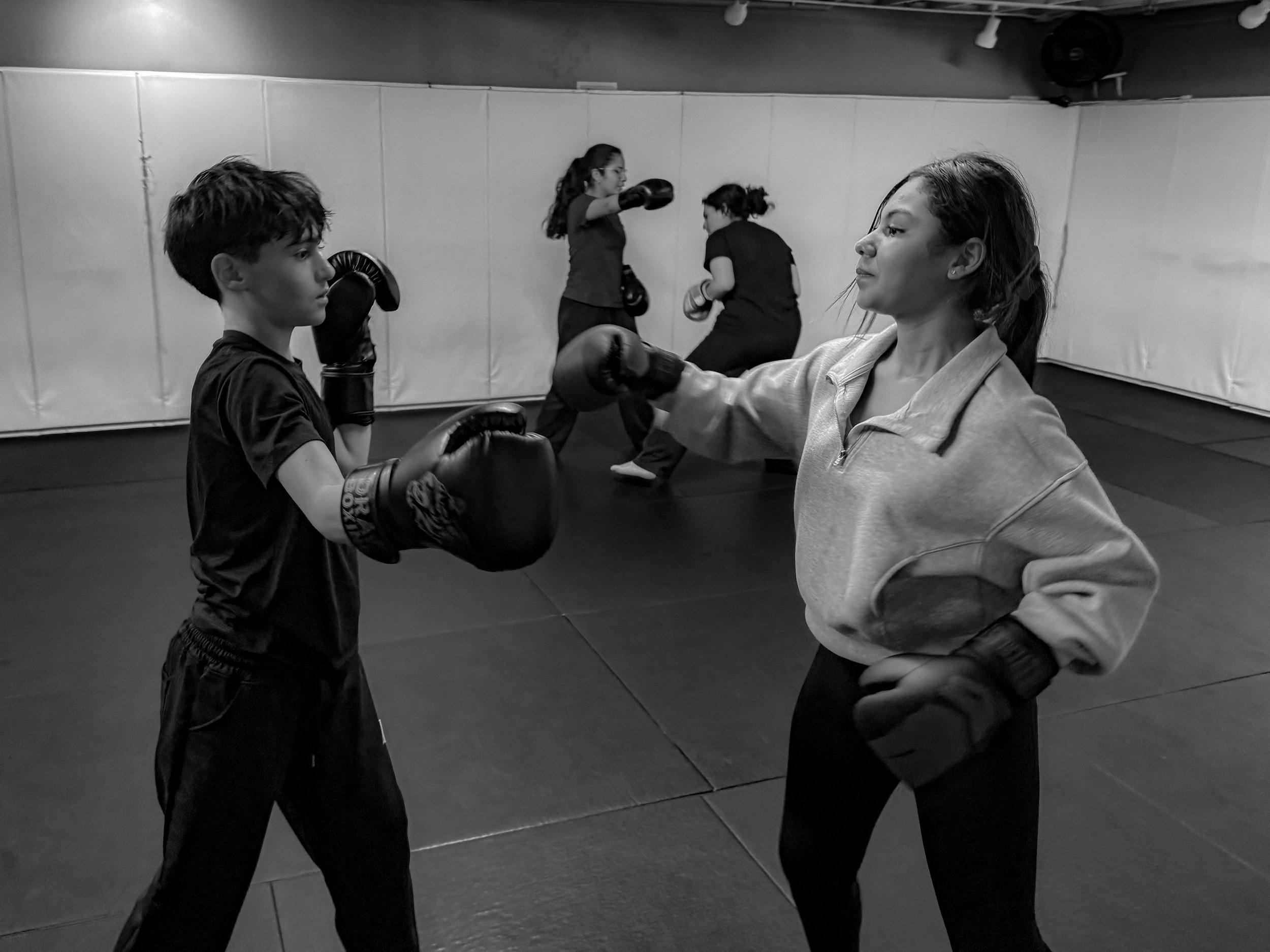 A woman and a young boy practicing boxing in a gym. The woman is wearing boxing gloves and the boy is wearing boxing gloves as well. They are facing each other in a fighting stance, with other people practicing boxing in the background.