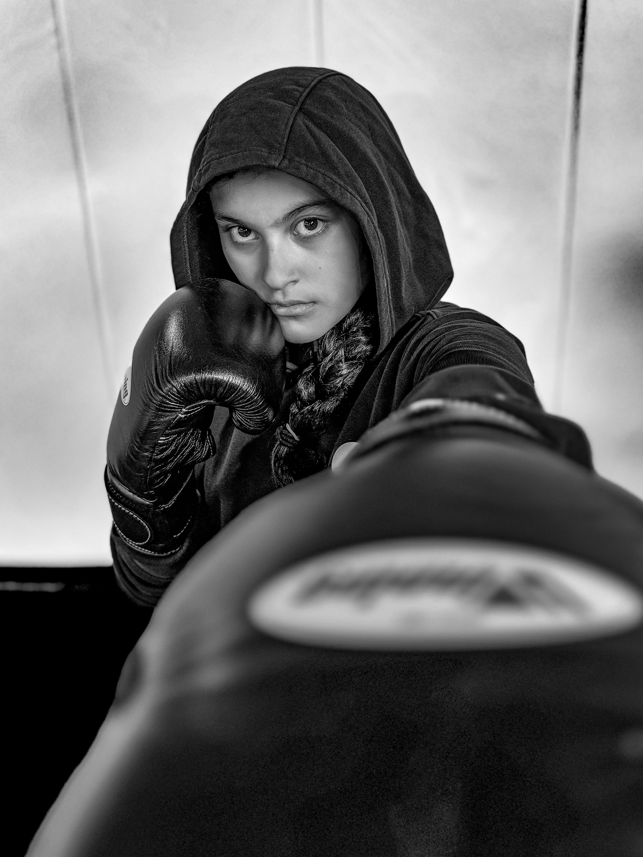 A young woman in a hoodie and boxing gloves taking a selfie with a camera, with the camera lens prominently in the foreground.