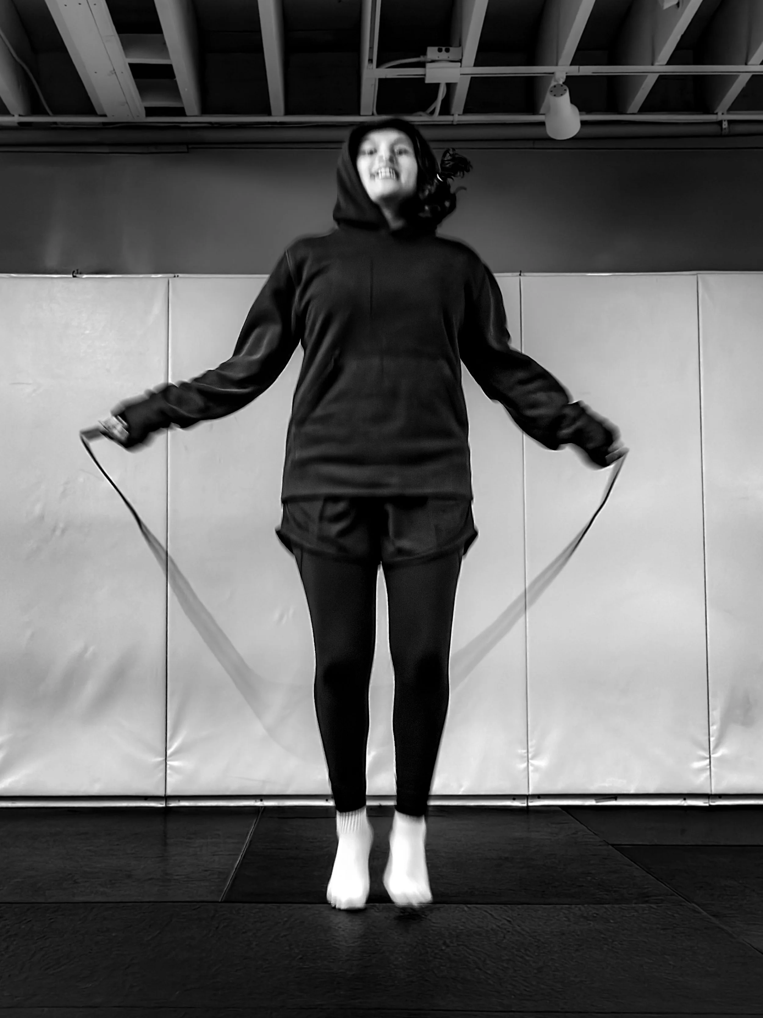 Black and white photo of a woman jumping with a jump rope indoors, wearing a hoodie and sweatpants, with a ceiling and plain wall in the background.