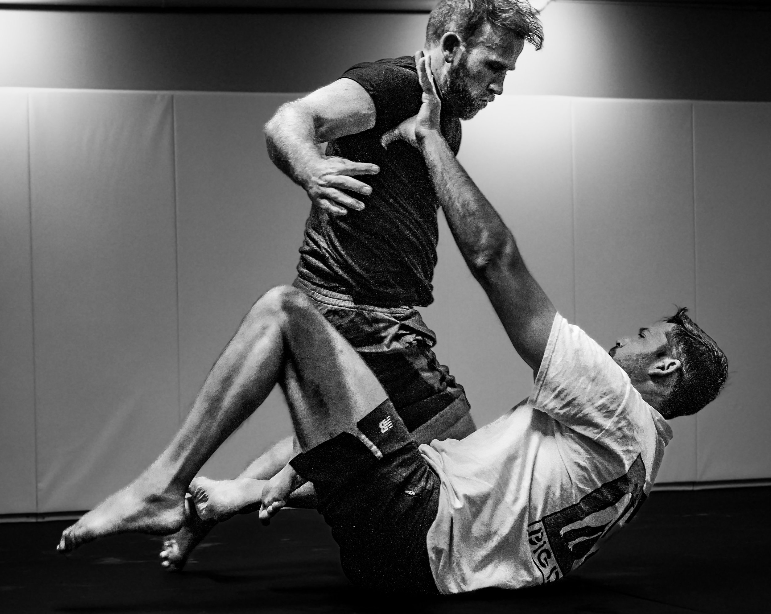 Black and white photo of two men practicing Brazilian Jiu-Jitsu on mats, one wearing a gi and the other in athletic clothes, engaged in a grappling move.