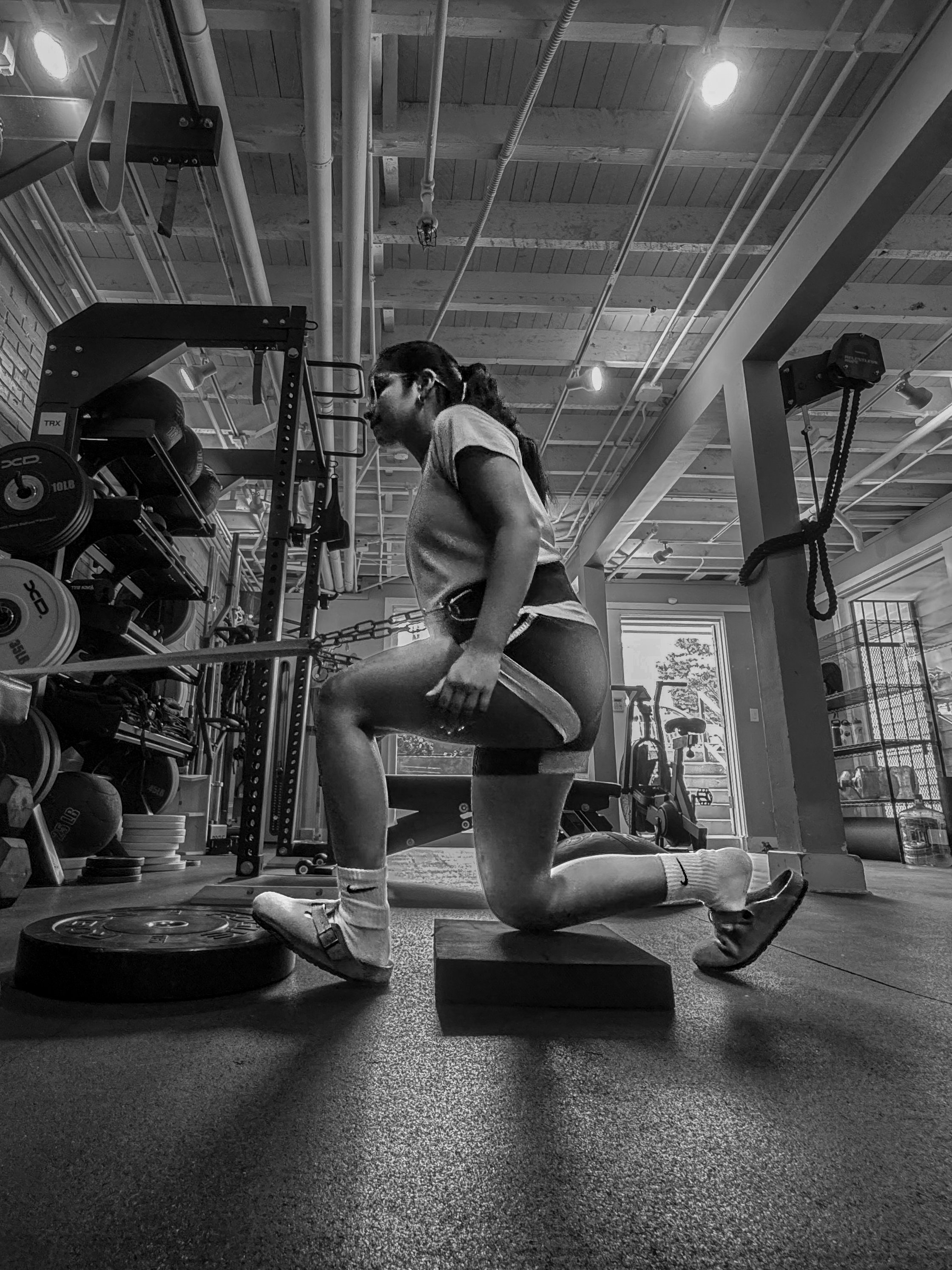 A woman in workout attire, including a t-shirt, shorts, and sneakers, performing a kneeling exercise with a weight plate on her thigh in a gym with weightlifting equipment and exposed ceiling pipes.