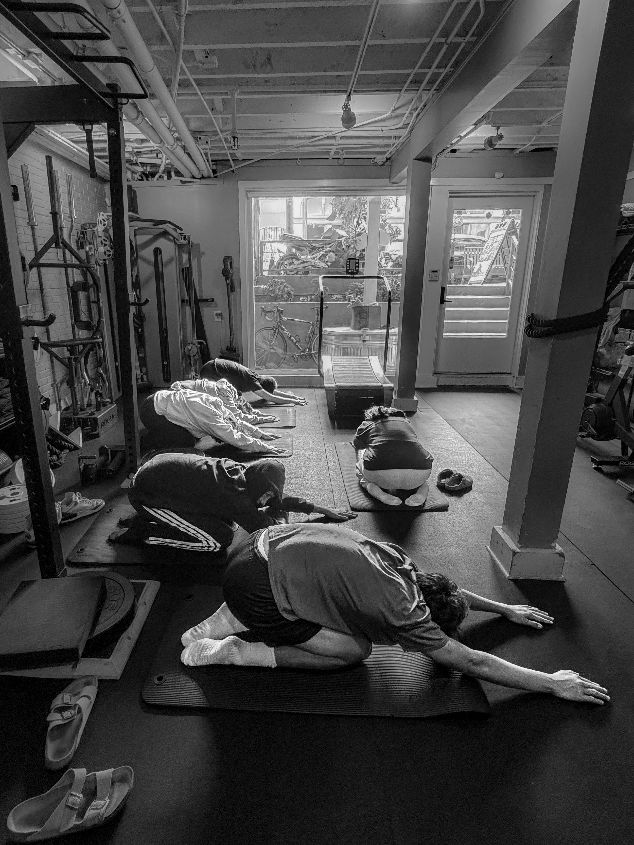 People participating in a yoga class on mats in a bright, industrial-style room with large windows and bicycles outside.