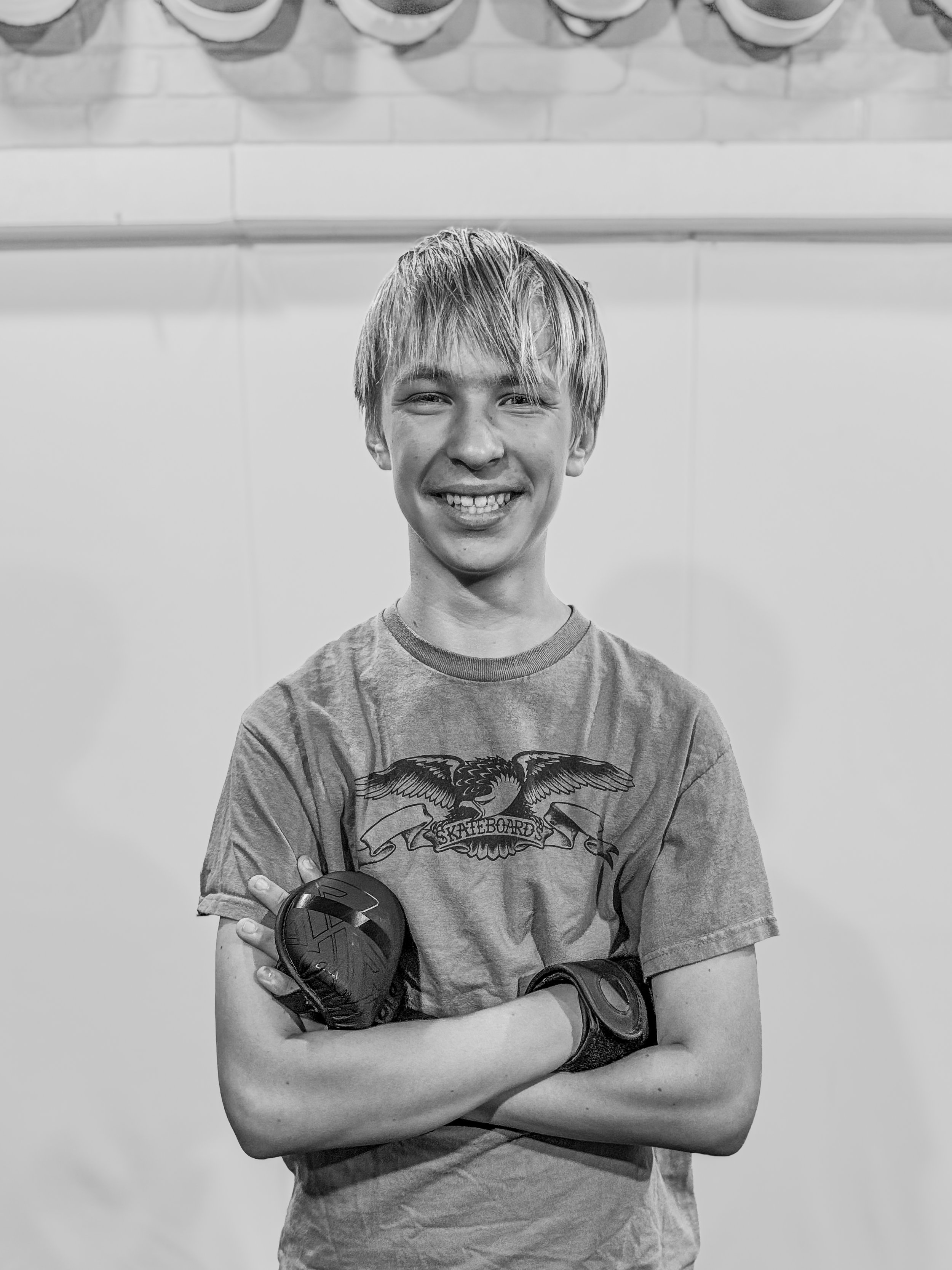A smiling teenage boy with blond hair, crossed arms, wearing a graphic t-shirt and wristwatches, standing against a plain wall with a row of plates or bowls on a shelf above his head.