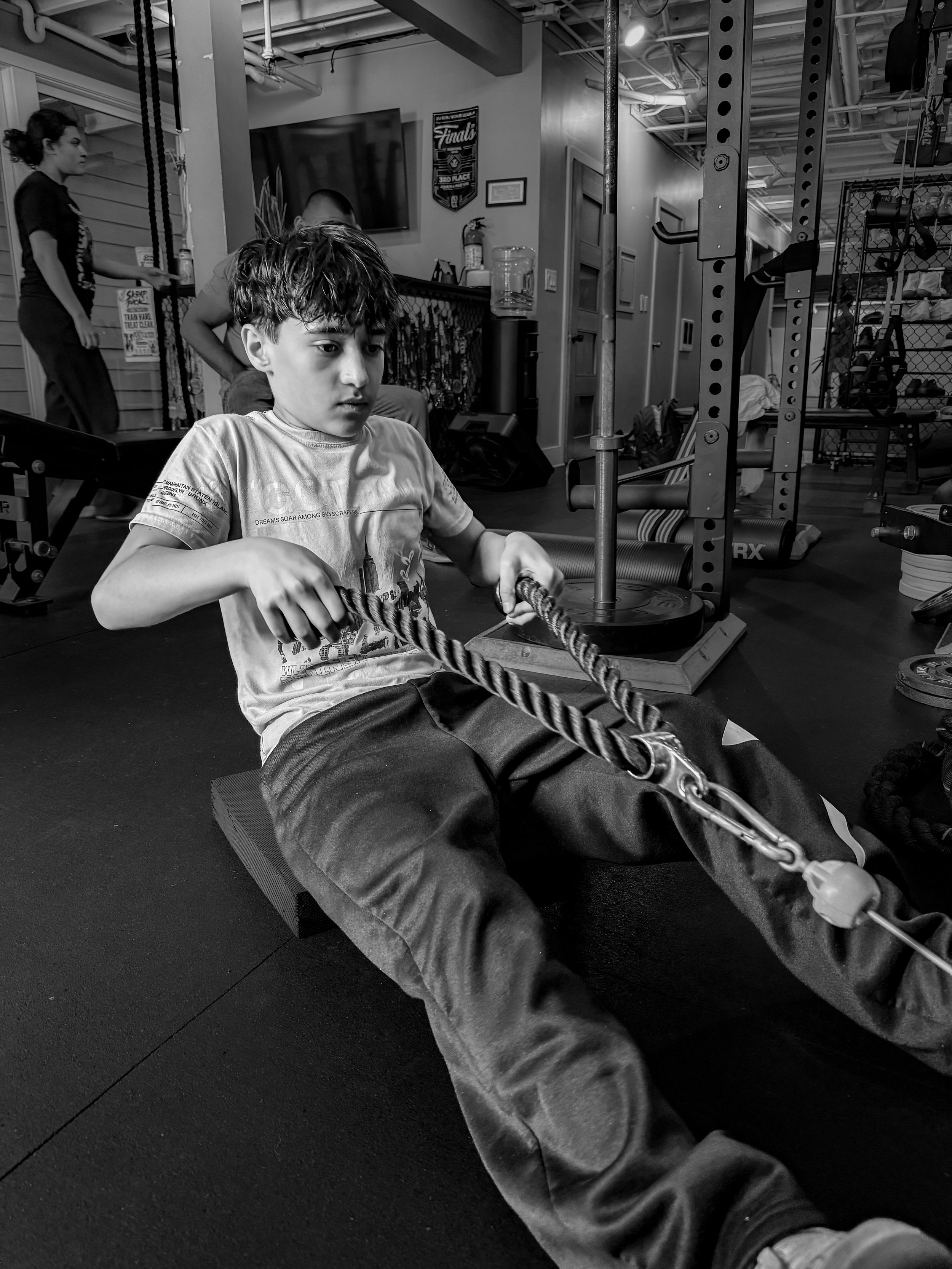 A young boy with curly hair sitting on the floor in a gym, holding onto a rope, with gym equipment and a woman in the background.