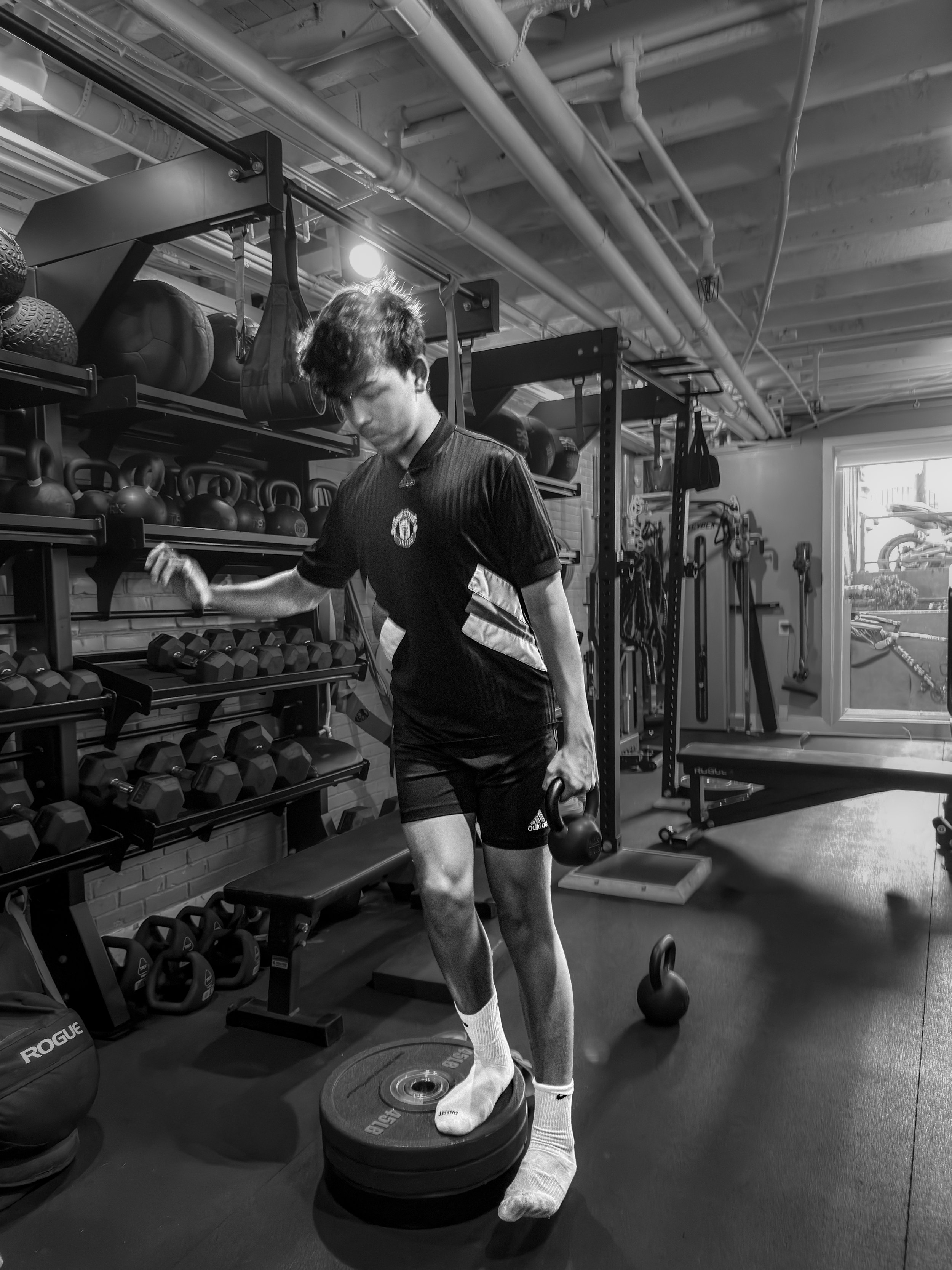 A person in a gym, lifting weights with a kettlebell in each hand while standing on a weight plate, with shelves of kettlebells and medicine balls in the background.