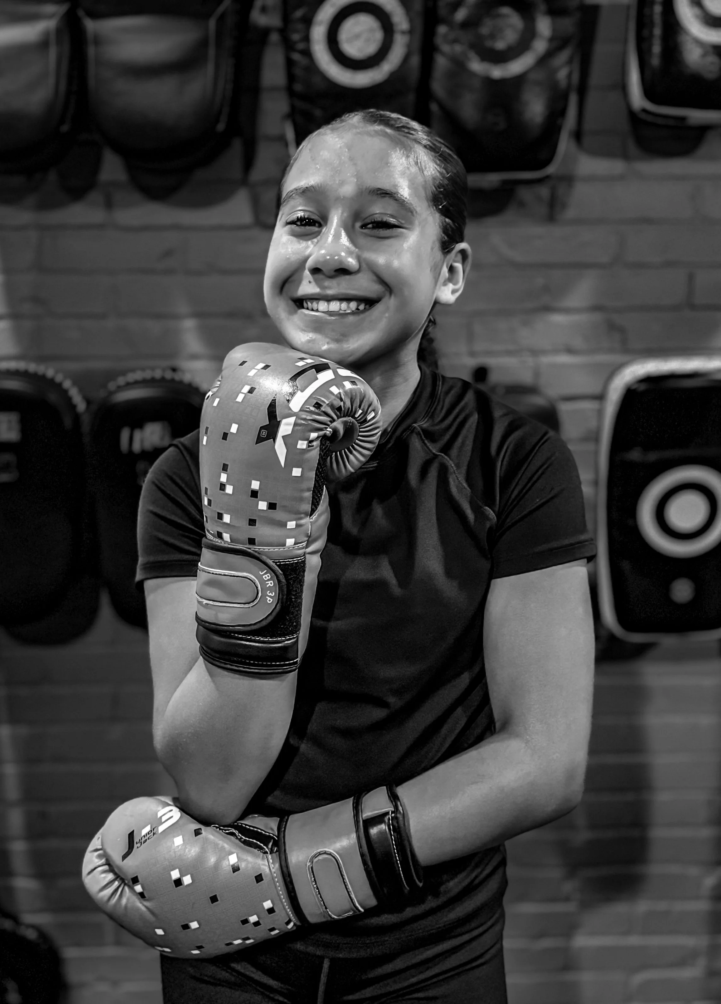 A young girl with a big smile, wearing boxing gloves, in a gym with a brick wall and punching bags in the background.