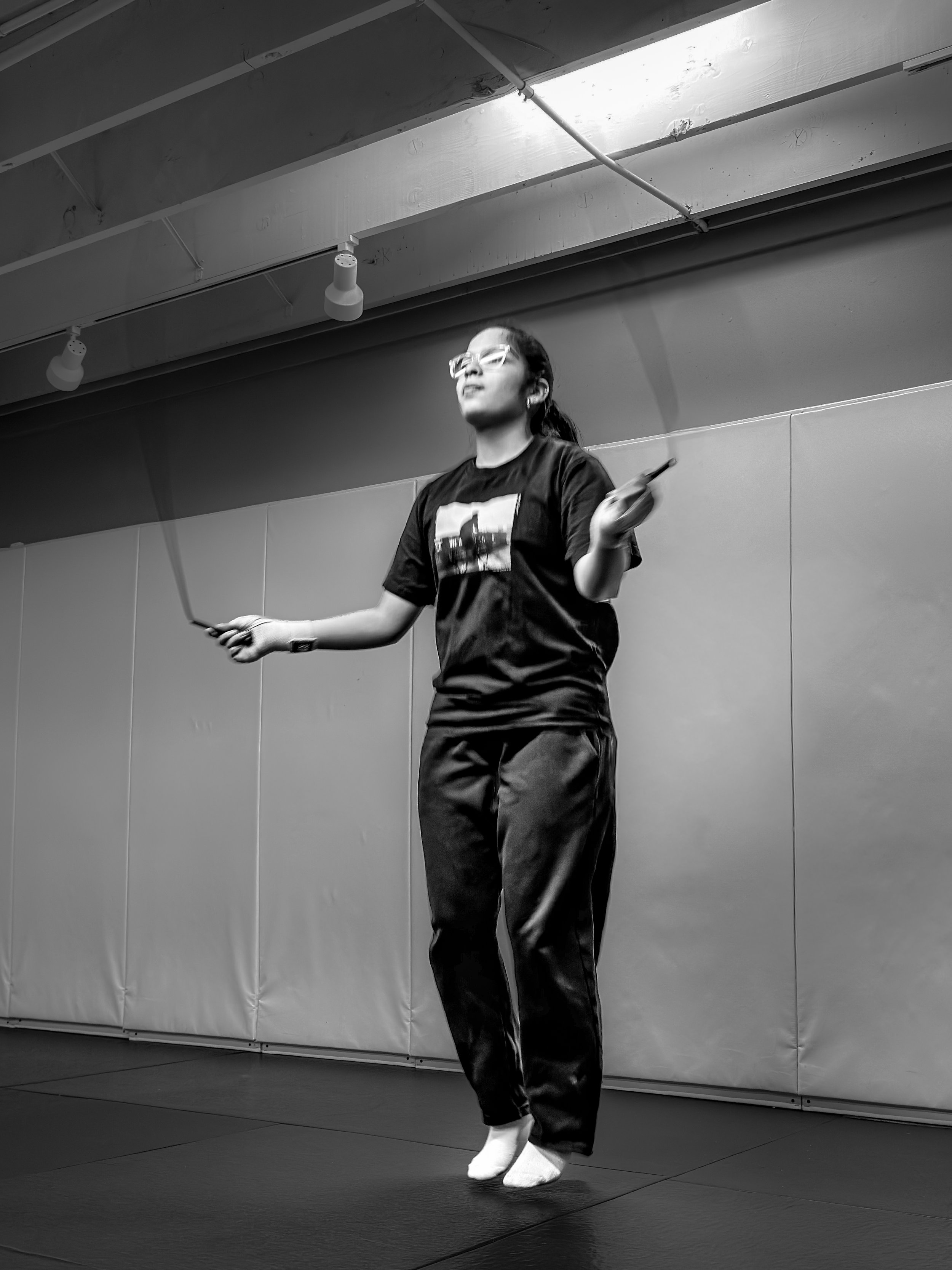 A woman with glasses and tied-back hair is performing a dance or exercise in a studio, holding a stick in each hand, dressed in a T-shirt and loose pants, wearing socks.