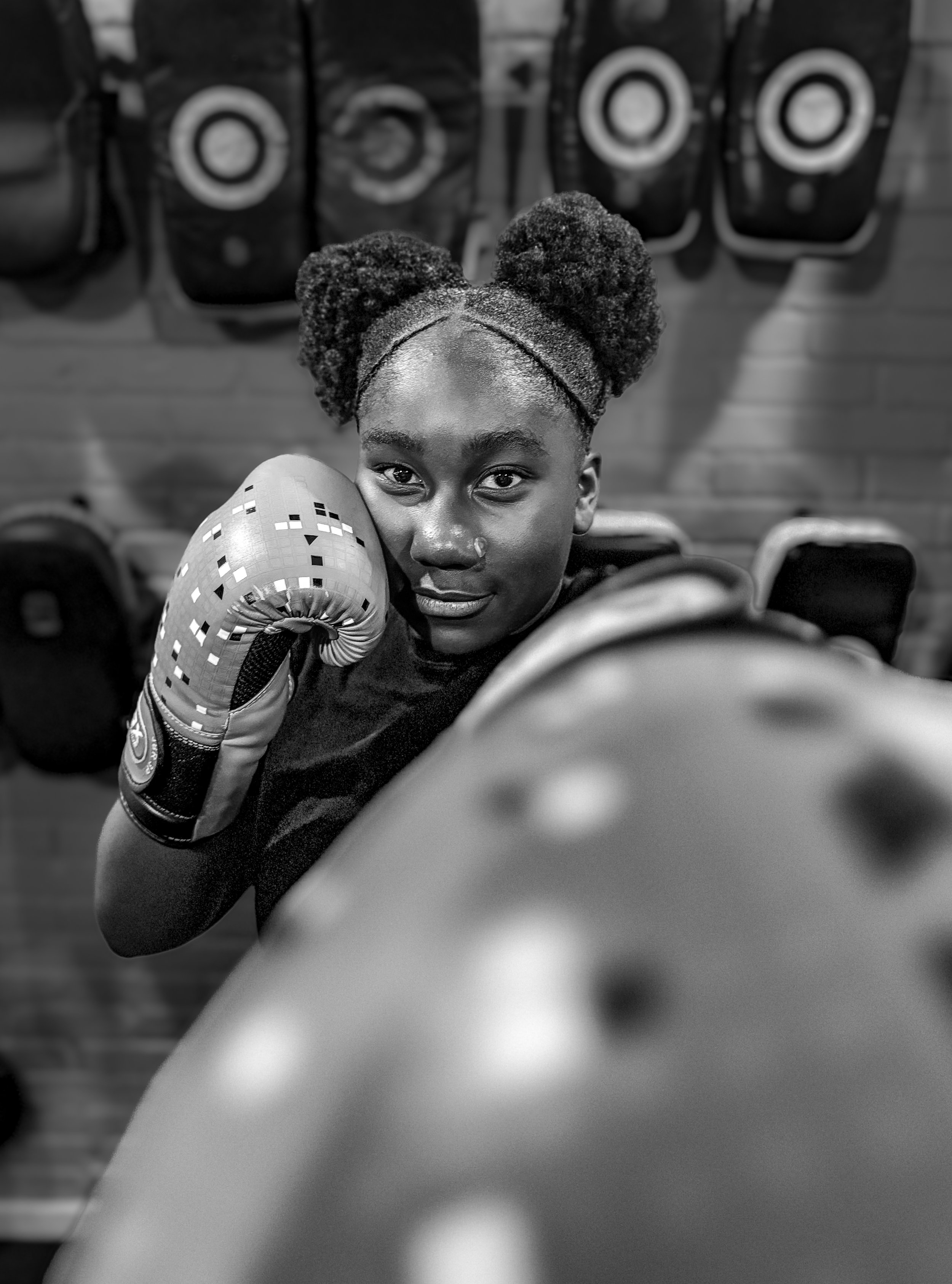 A young woman with styled hair in buns and wearing boxing gloves, posing in a boxing gym with punching bags in the background.