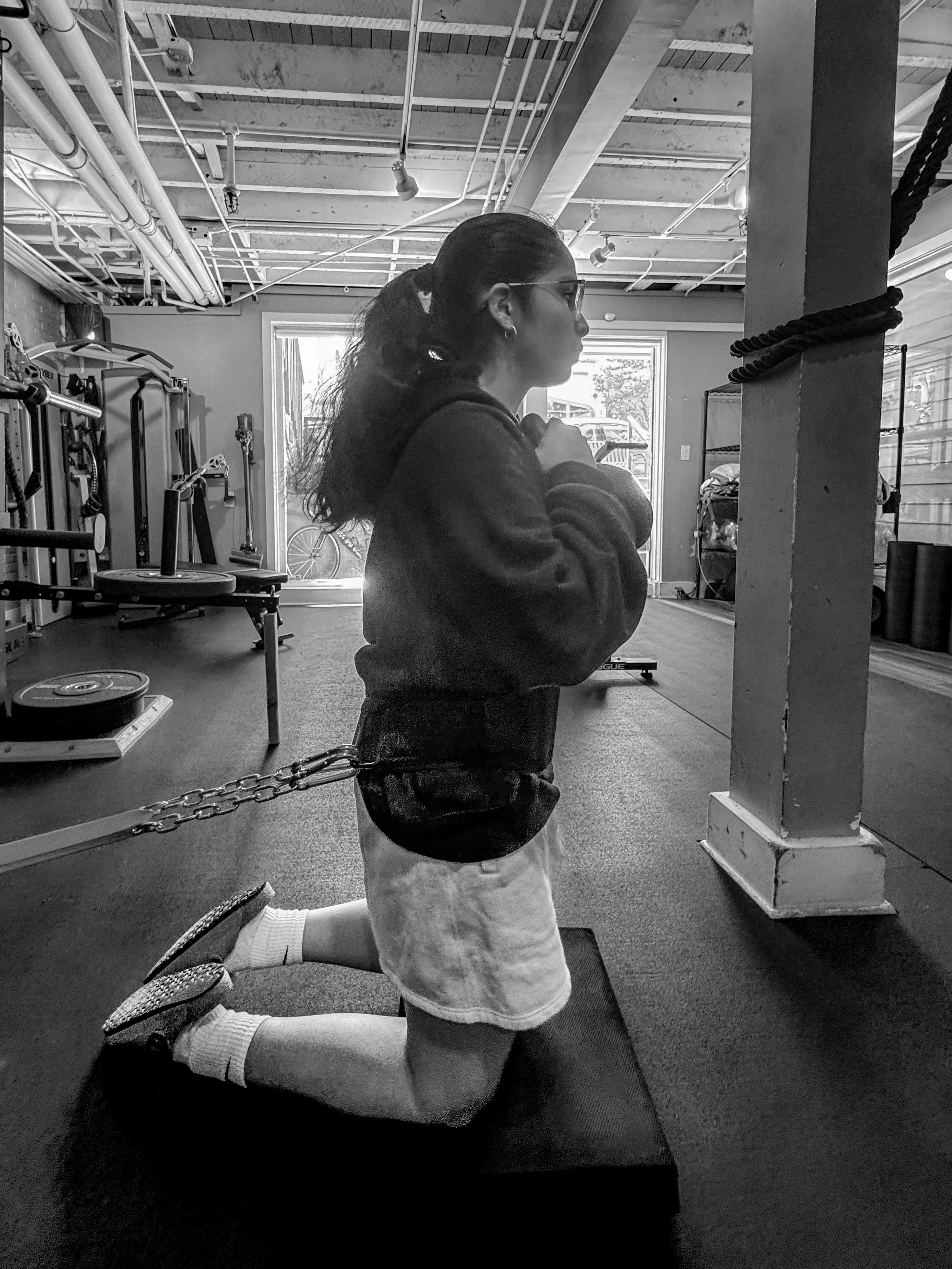 A woman kneeling on her right knee inside a gym, wearing athletic clothes, with her left hand on her chest, looking straight ahead. The gym has exercise equipment on the left side and a large window in the background.