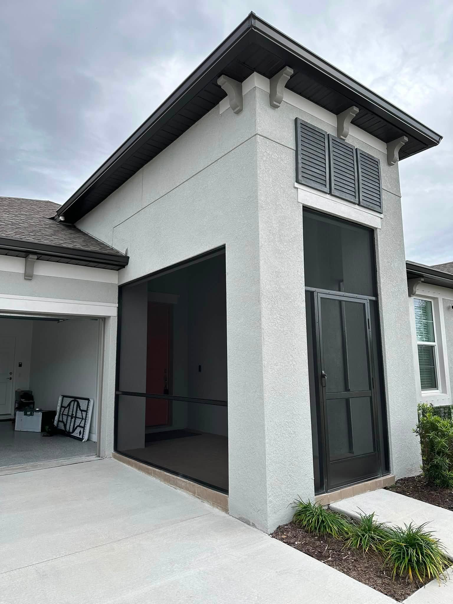 The corner of a modern house with a white stucco exterior, black window shutters, and a screened porch. The house has a dark gray roof with overhanging eaves, and there are small plants and a concrete patio in the yard.