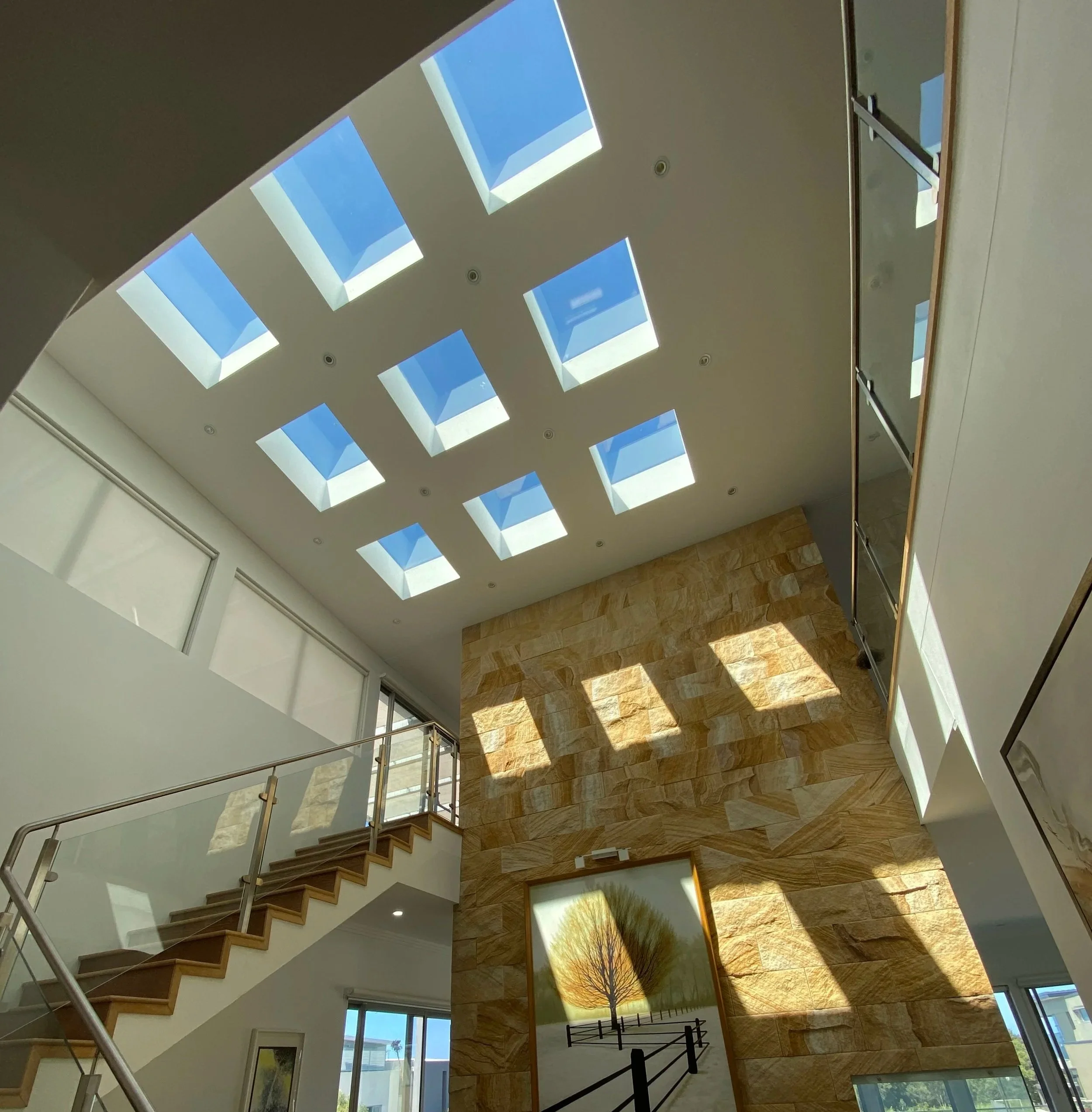 Interior view of a modern house with large skylight windows letting in natural sunlight, wooden staircase, artwork, and railing.