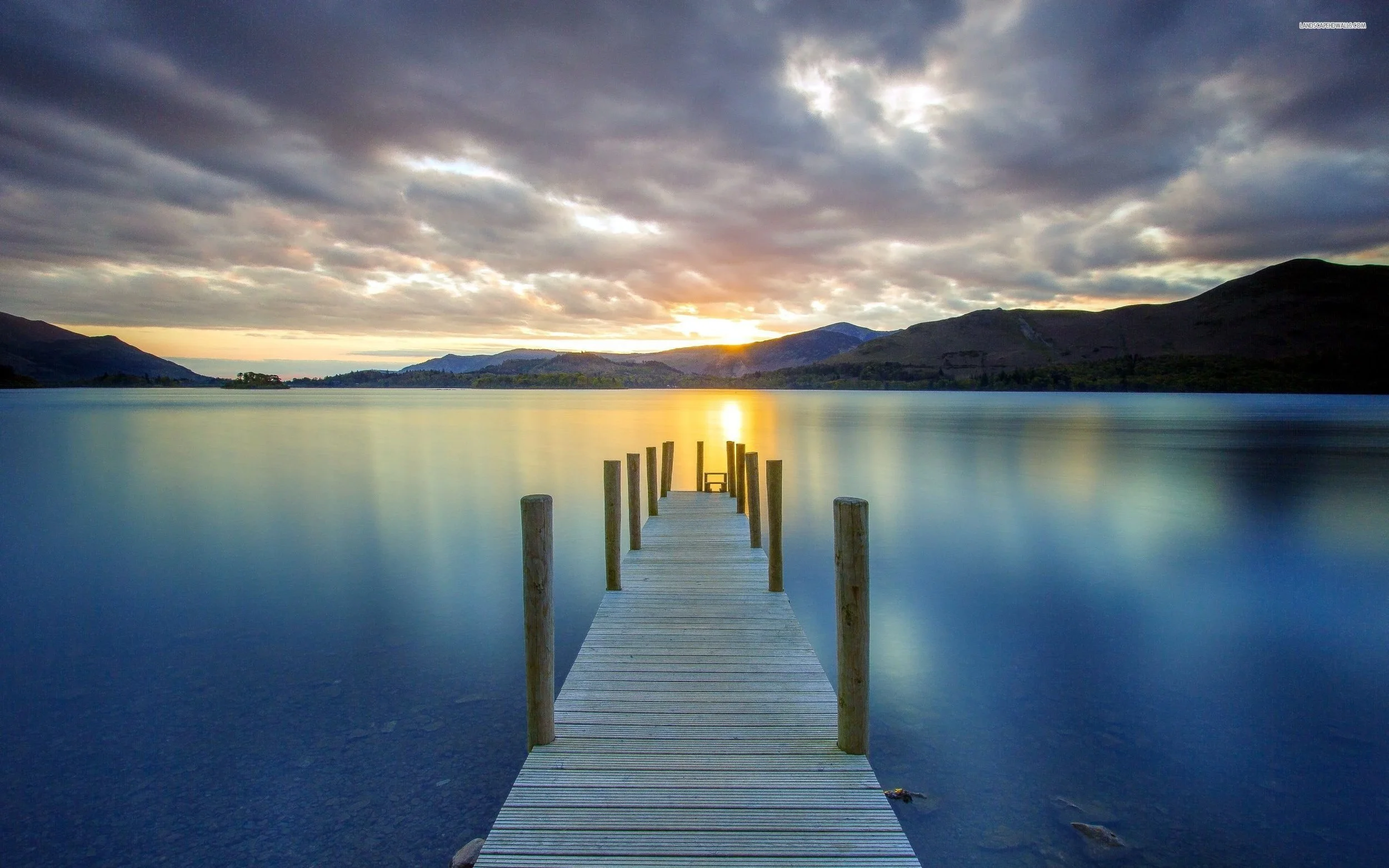 A wooden dock extends into a calm lake during sunset, with mountains and cloudy sky in the background.