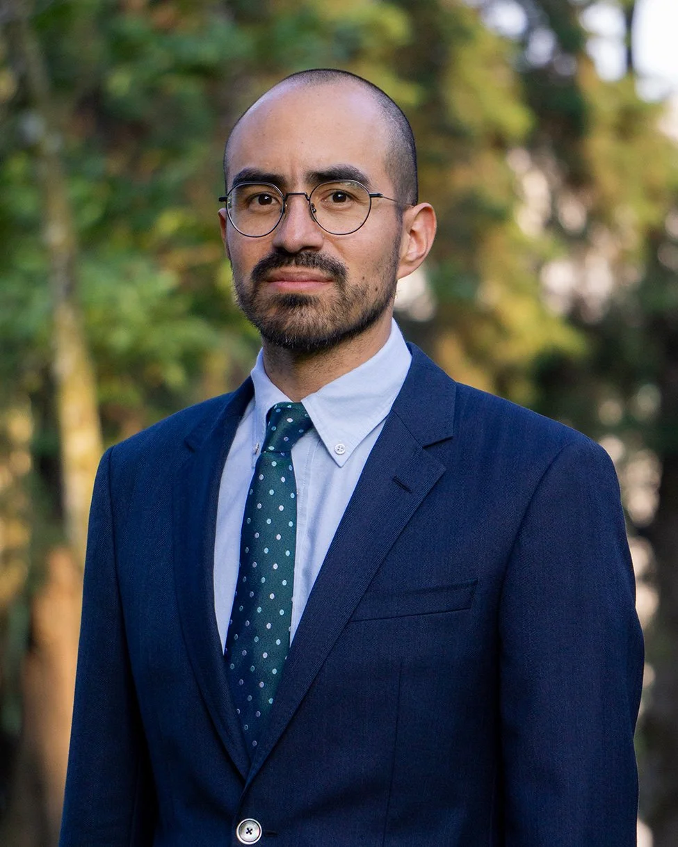 A man with glasses, a beard, and a receding hairline wearing a navy suit, light blue shirt, and a polka dot tie, standing outdoors with blurred trees in the background.