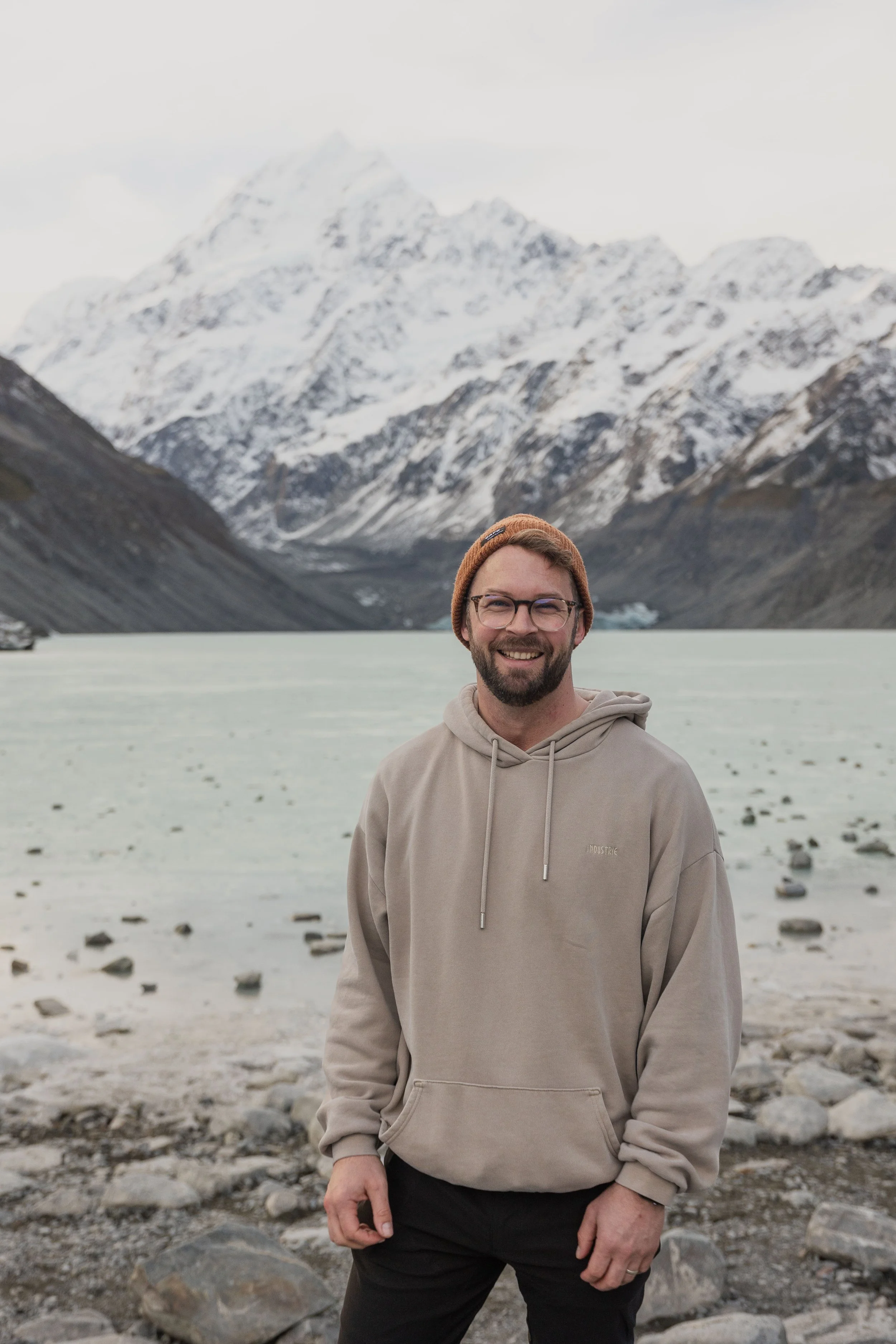 A man in a beige hoodie and black pants standing on a rocky shore with a snow-capped mountain range and icy lake in the background.