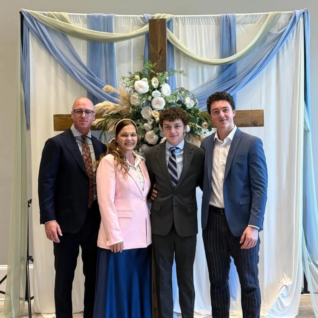 A family of four standing in front of a floral cross backdrop at a church or event. The adults are dressed in formal attire, with the man in a dark suit and the woman in a pink blazer and blue skirt. The two young men are also in suits, one in a gray suit with a striped tie and the other in a blue suit with pinstripe pants.