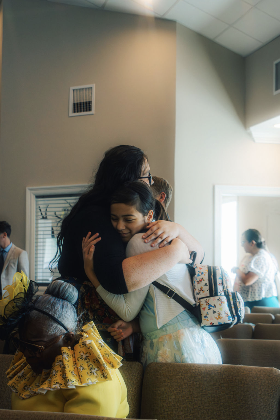 Two women hugging warmly in a room with other people, some sitting and some standing, during a gathering or celebration.