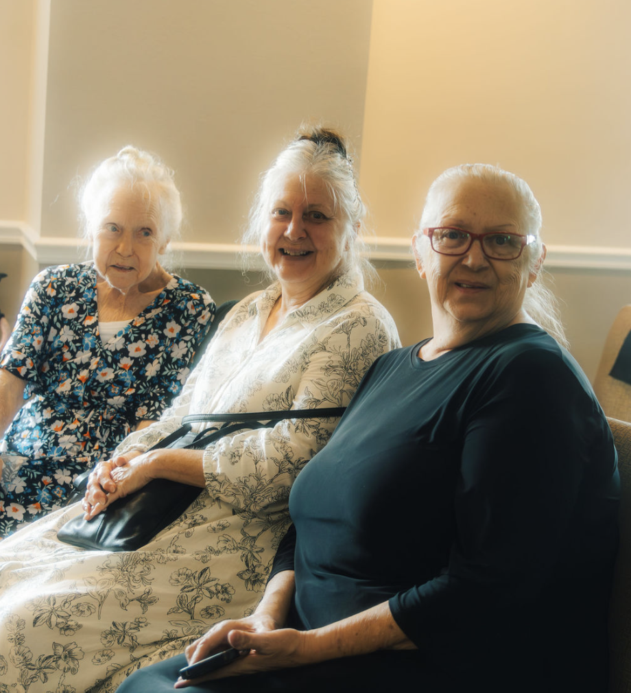 Three elderly women sitting together, smiling, in a room with beige walls.