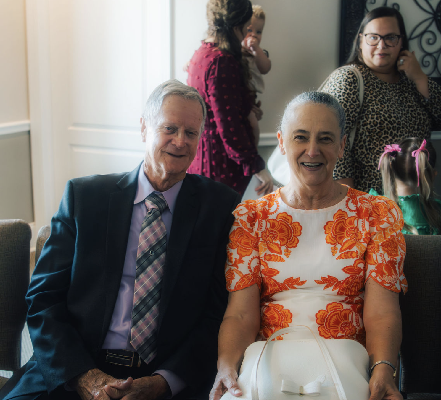 An elderly man in a suit and an elderly woman in a floral dress sitting together at a gathering, smiling, with other people and children in the background.