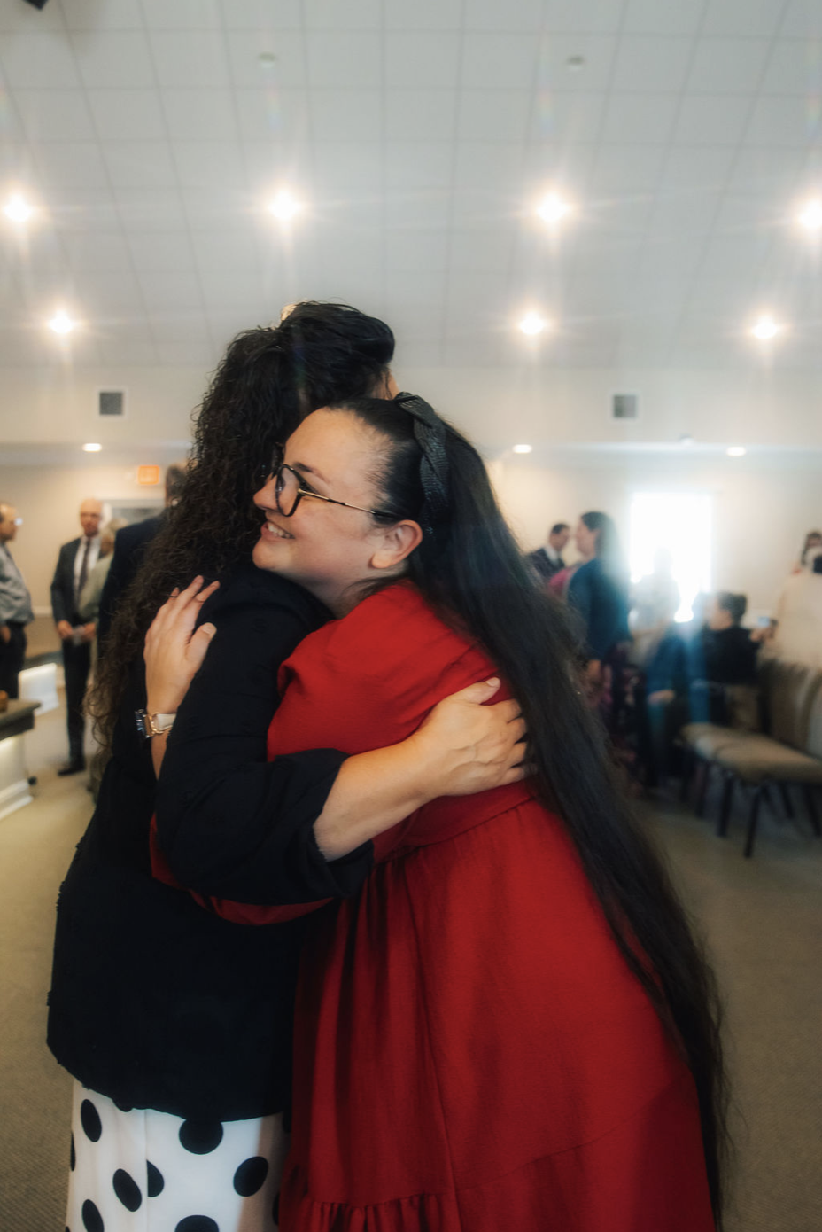 Two women hugging and smiling in a room filled with other people, with a brightly lit ceiling.