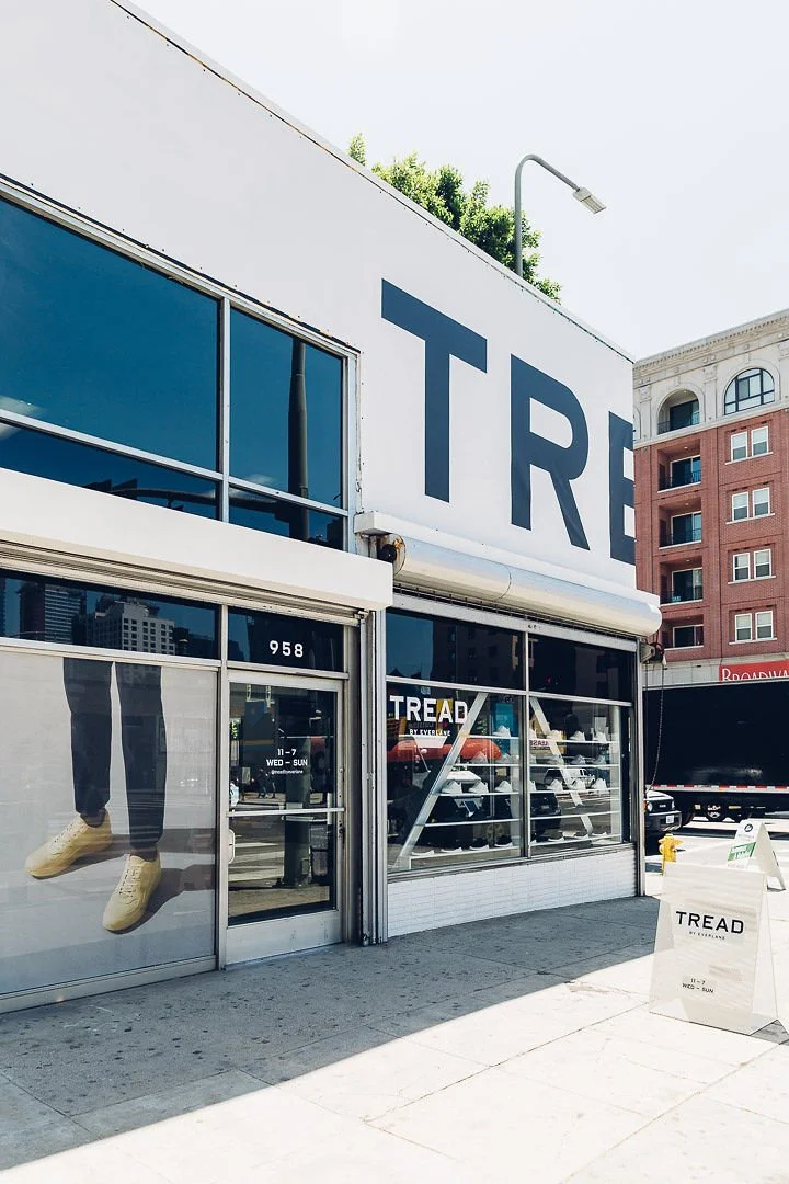 Outside view of a TREAD store with large glass windows displaying sneakers, a sign with store hours, and a sidewalk in front.