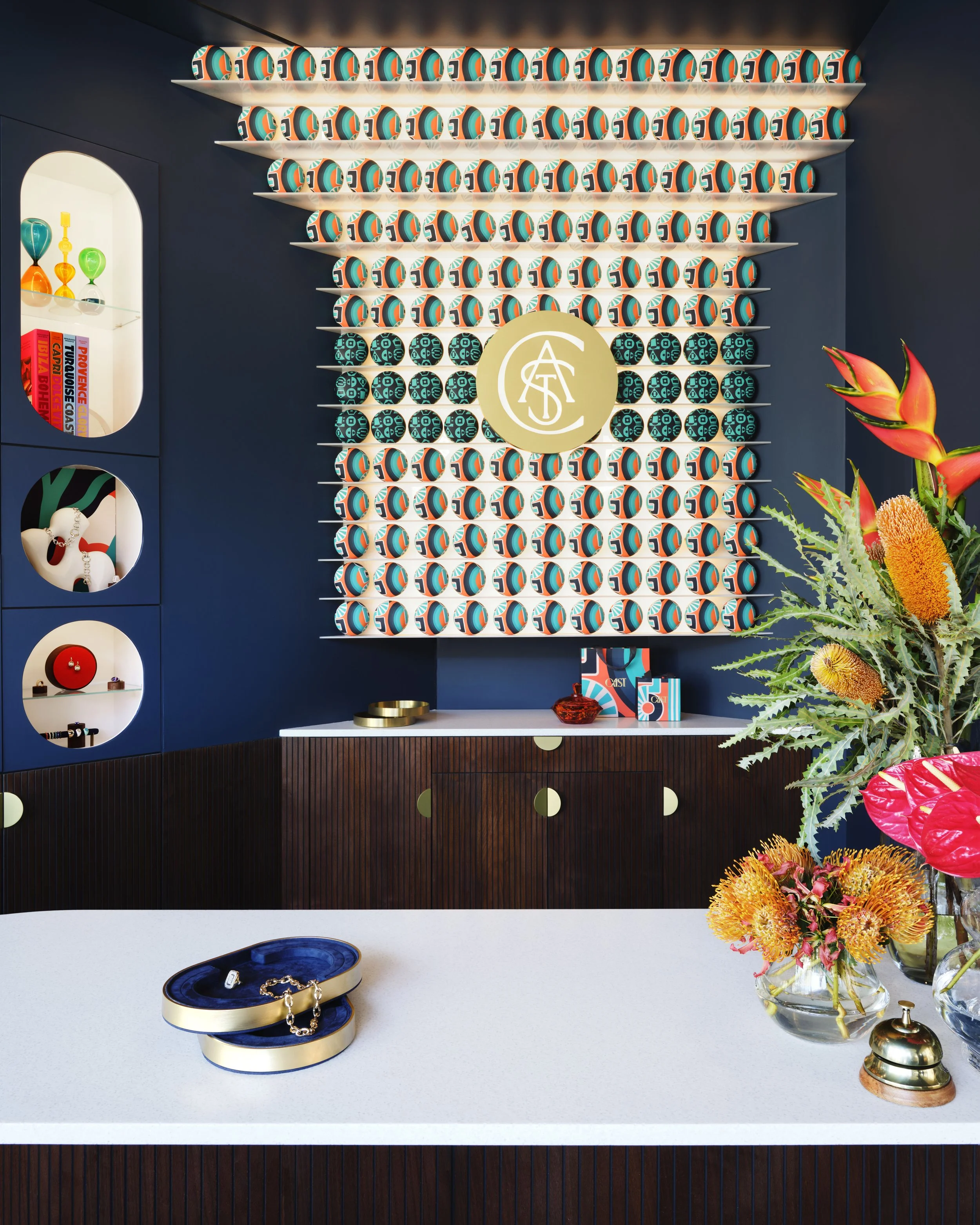 View of a jewelry display table with an open jewelry box containing a gold bracelet on a white surface. To the right, a glass vase with a bouquet of colorful flowers including pink, orange, and yellow blooms and green foliage. In the background, a dark blue wall with white circular shelves holding decorative items such as glass vases, books, and jewelry. Prominent round golden logo with white lettering in the center of a patterned display of circular objects.