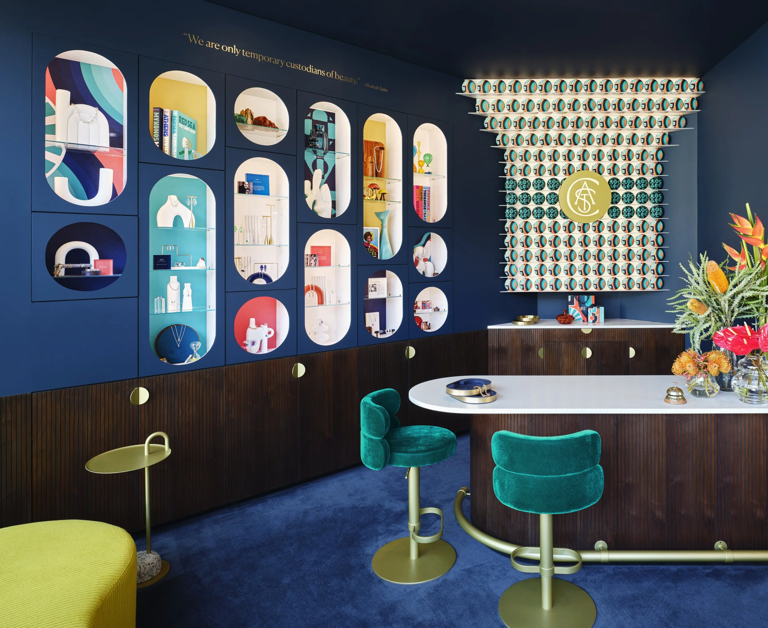 A stylish interior with a dark blue wall featuring illuminated display shelves with various decorative items, and a wall decoration made of vertically stacked colorful patterned tubes. There are two teal velvet chairs with curved metal legs and a white round table with accessories on it. A green upholstered seat and a yellow side table are also visible, along with a floral arrangement in the foreground.