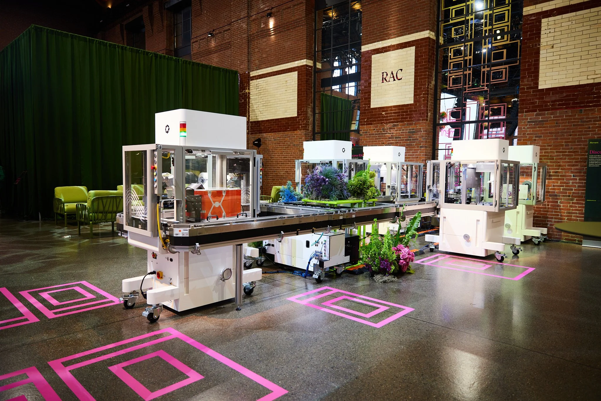 Exhibition space with robotic machinery and greenery displays, pink floor markings, brick wall with green curtain backdrop, and large windows showing an industrial interior.