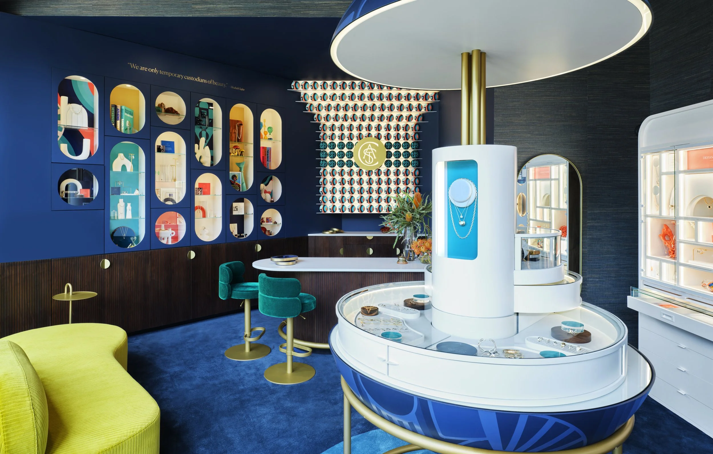 Interior of a modern jewelry store with vibrant blue walls, display cases filled with jewelry, and colorful seating including a yellow sofa and teal chairs. There is decorative wall art and a quote on the wall that says, "We are only temporary custodians of beauty."