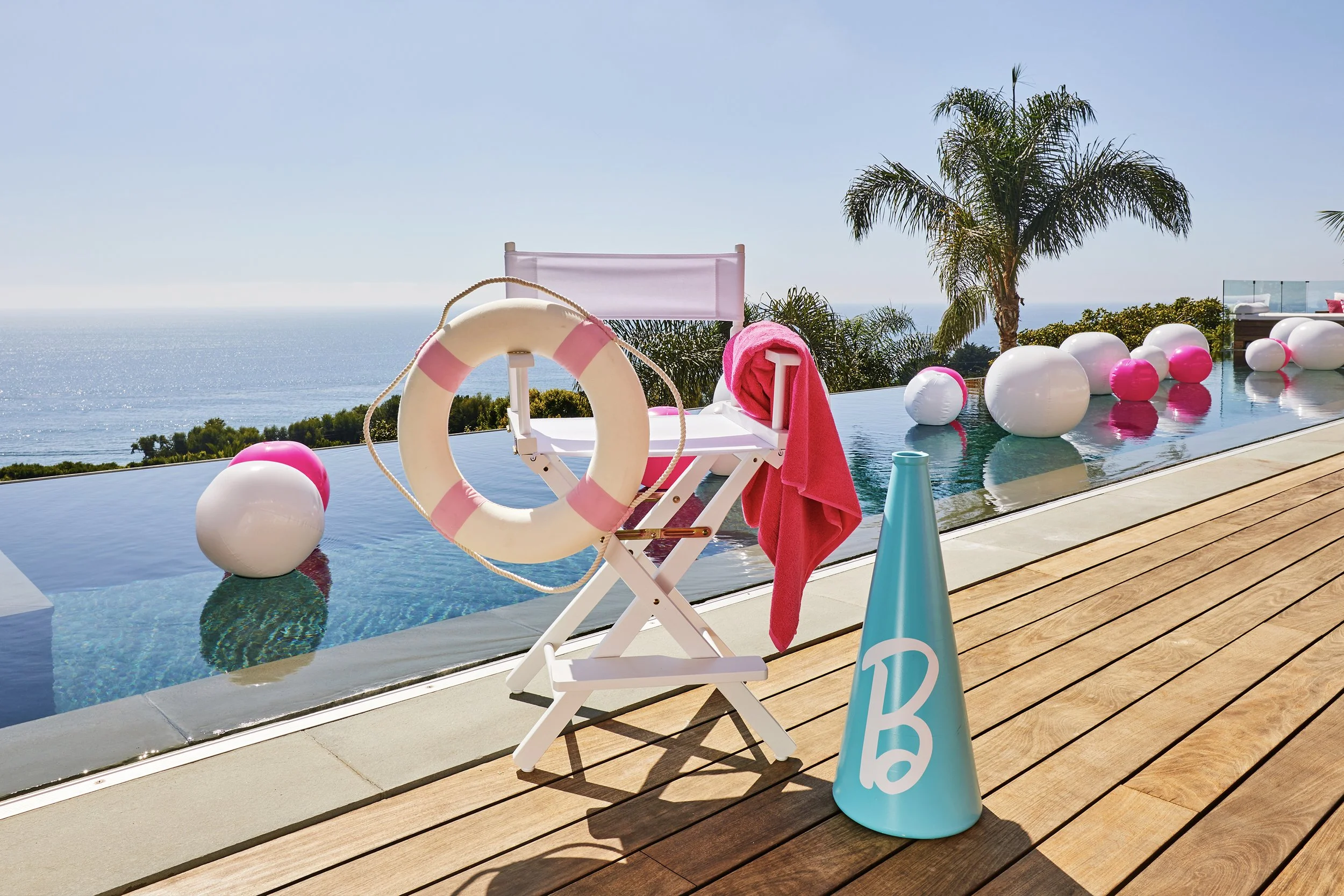 Poolside scene with pink and white balloons, a lifebuoy, a pink towel, and a folding chair near a pool, palm trees, and ocean in the background with clear blue sky.