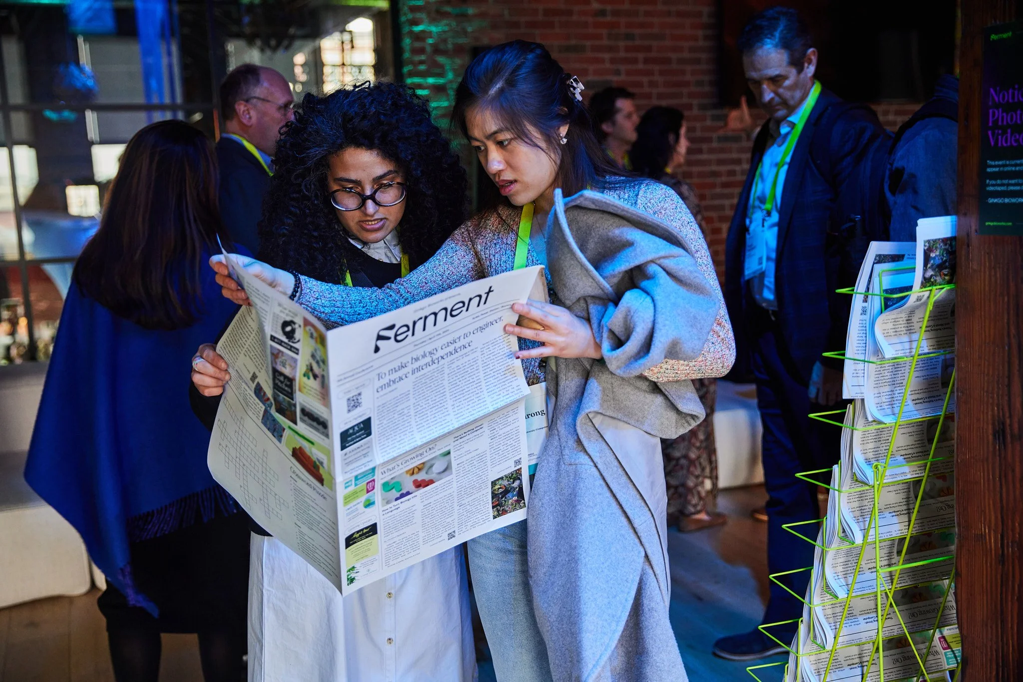 Two women are reading a newspaper together at an indoor event, with other people in the background.