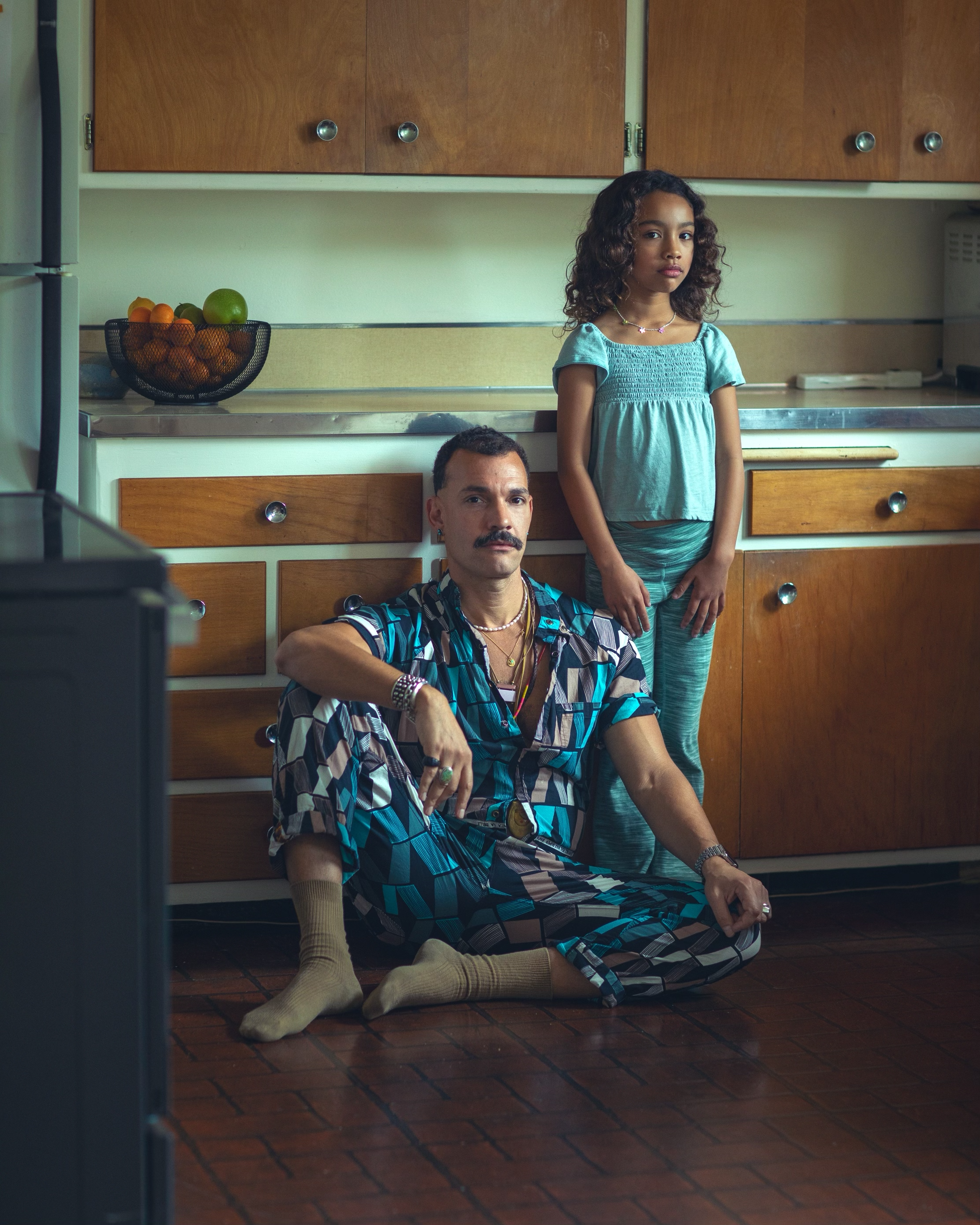 A man sitting on the floor with a young girl standing beside him. Both are in a kitchen with wooden cabinets and a fruit bowl on the counter. The man has a mustache and is wearing a patterned short-sleeve shirt with accessories. The girl has curly hair and wears a teal top with matching pants. They are looking toward the camera.