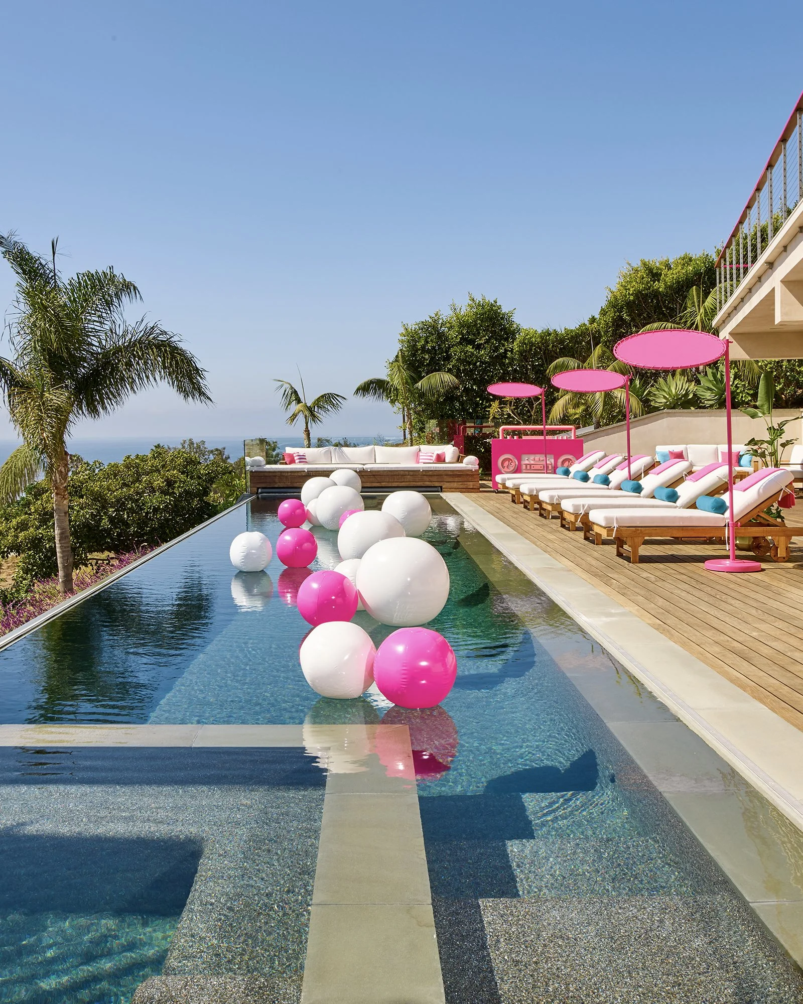 Luxury outdoor pool area with floating pink and white balloons, white lounge chairs with blue cushions, pink umbrellas, and lush greenery under a clear blue sky.