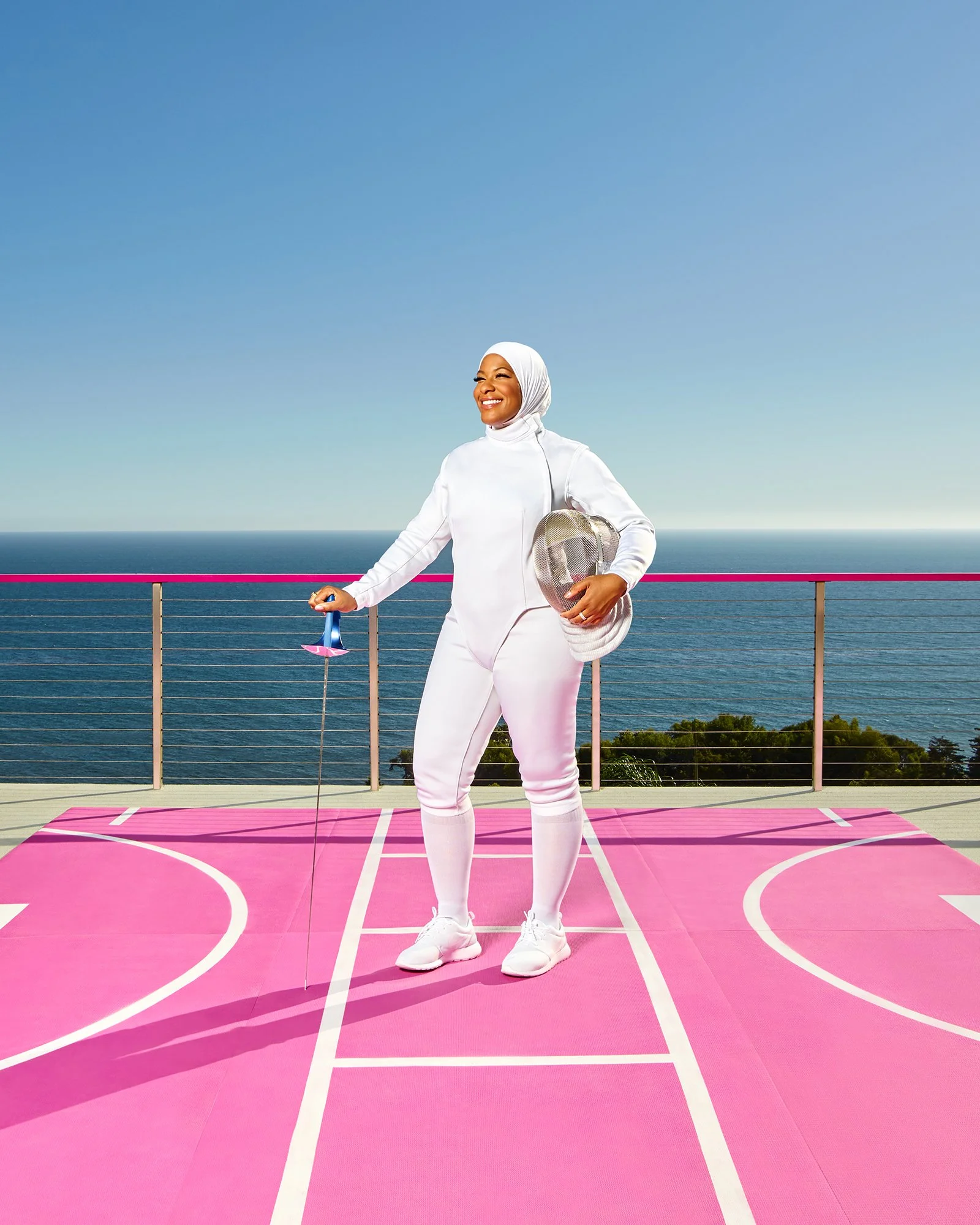 A woman wearing a white hijab and all white sportswear, holding a tennis racket and a paddle, standing on a pink outdoor basketball court with the ocean and a clear blue sky in the background.