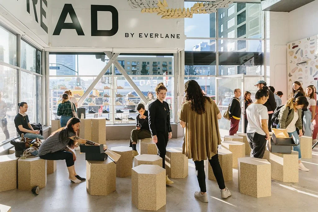 People shopping in a modern retail store with large windows, seating blocks, and a sign that reads "THE AD BY EVERLANE".