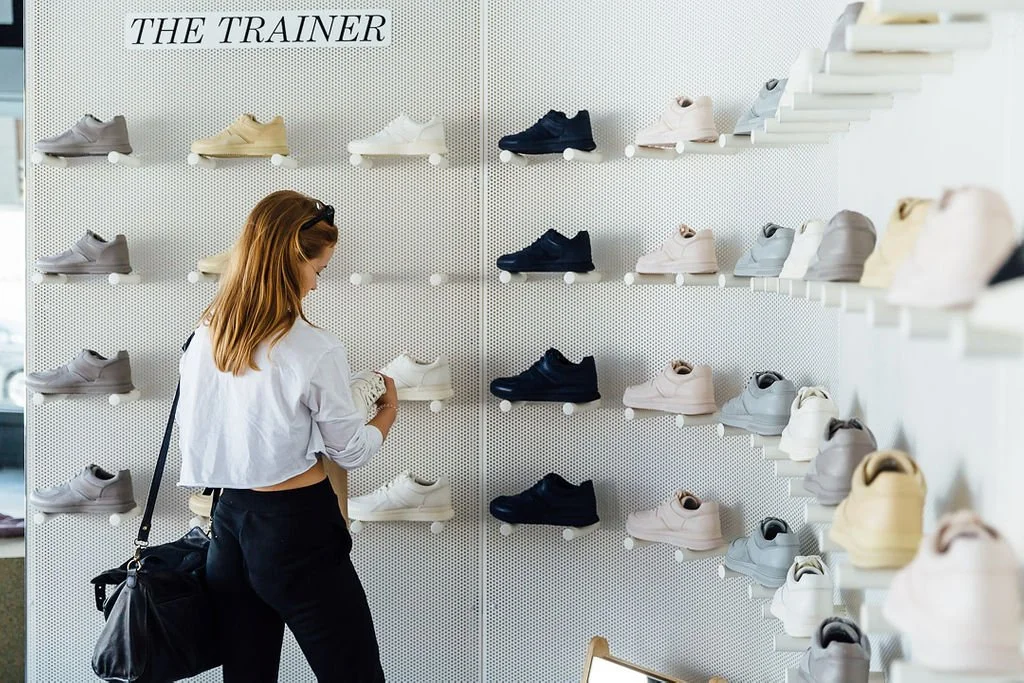 A woman shopping for shoes in a store, looking at various sneakers on display shelves.