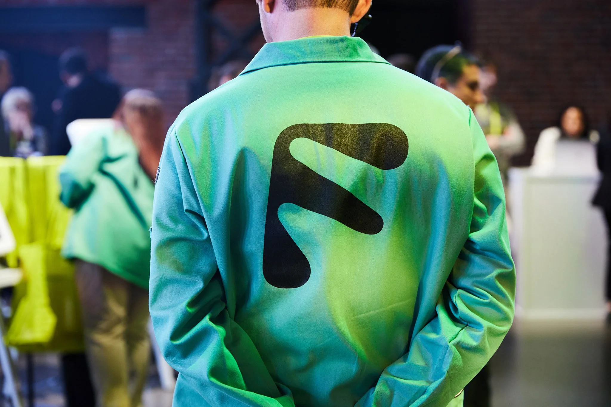 Close-up of a person wearing a light green jacket with black abstract designs, standing in a crowded indoor setting.