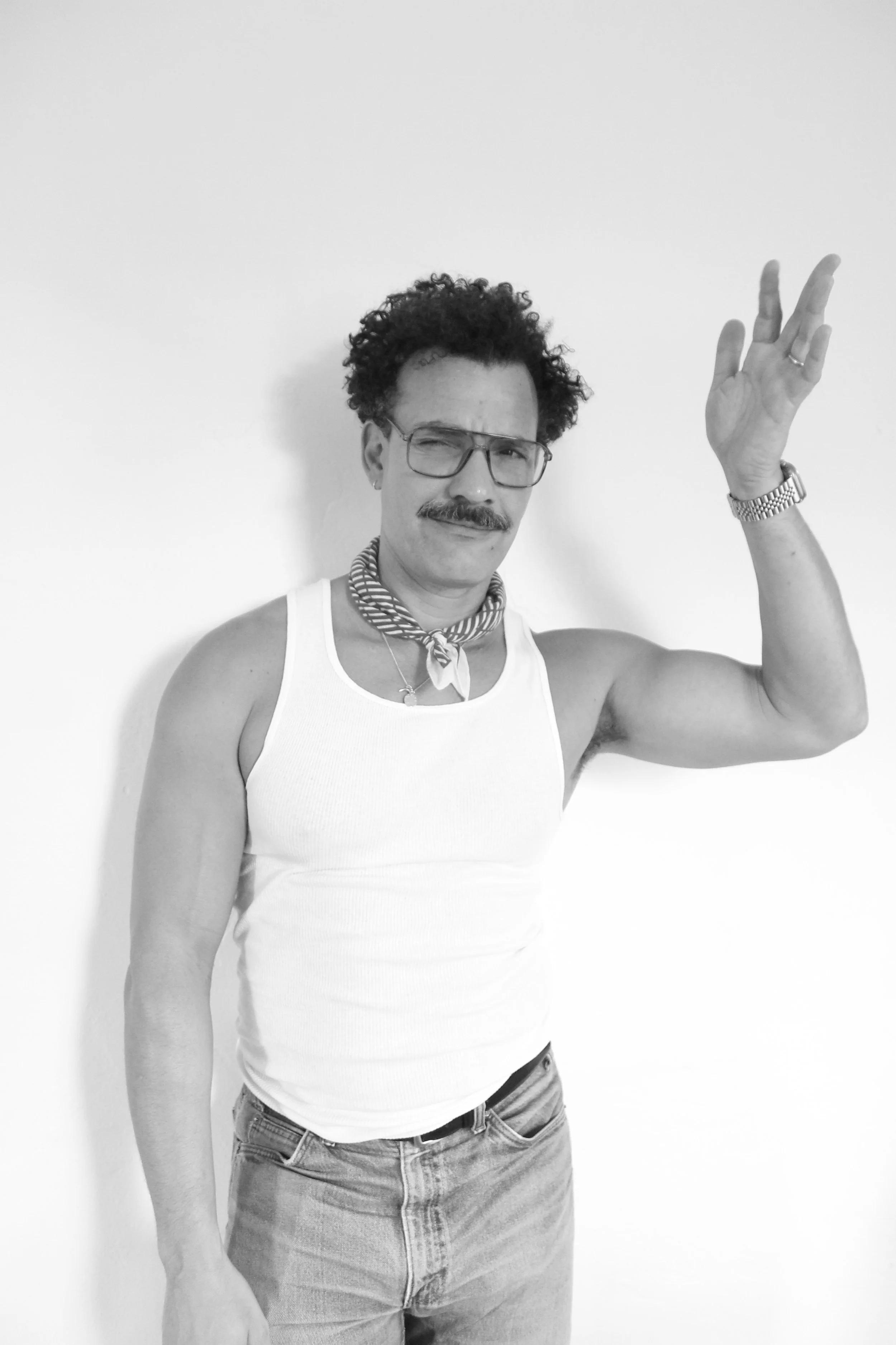 A person with curly hair, glasses, a mustache, wearing a white tank top, jeans, a striped scarf, and a watch, raising their right hand in front of a plain white wall.