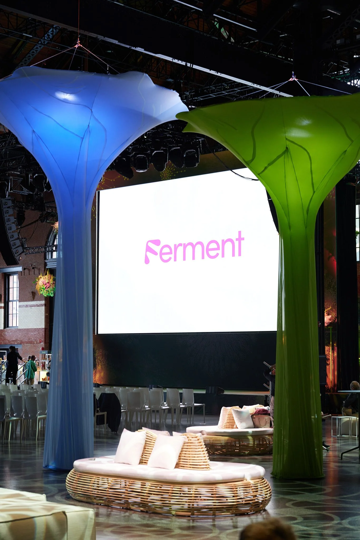 Indoor event space with large woven daybed with white cushions in front of a large white screen displaying the word 'Ferment' in pink. Two colorful fabric structures, one blue and one green, hang from the ceiling, creating a decorative arch. Chairs are arranged in rows facing the screen, and a few people are seen in the background.