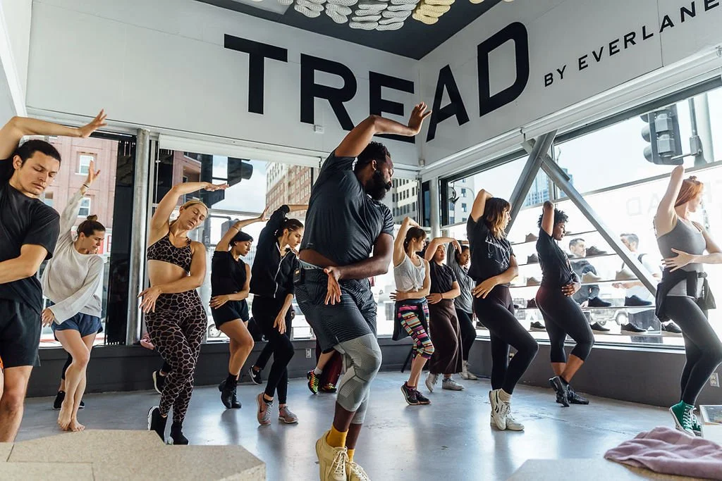 Group of diverse people participating in a dance or fitness class inside a modern studio with large windows, city buildings outside, and a 'TREAD by Everland' sign on the wall.