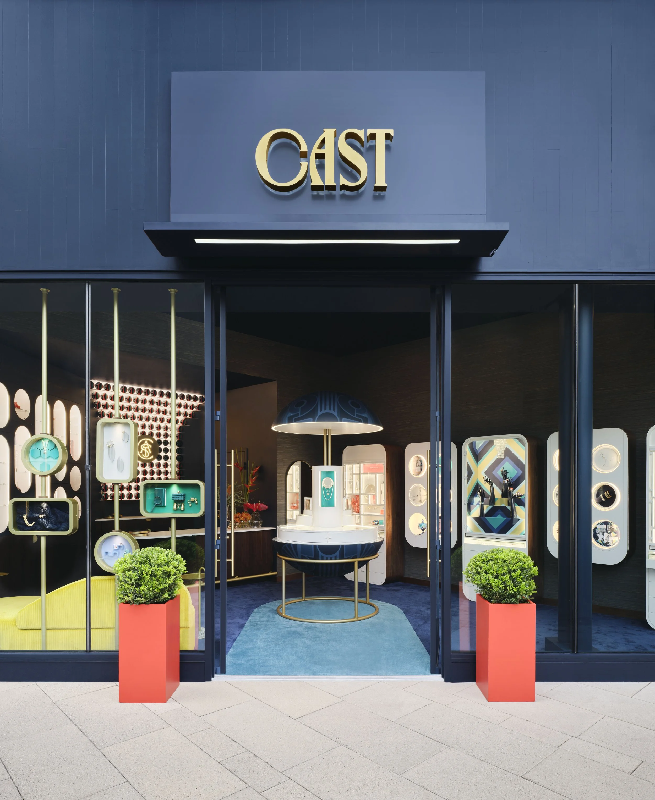 Storefront with a sign that says 'GAST' above the entrance. The interior is visible through large glass windows, showing shelves with jewelry and decorative items. Two potted green plants in red planters are positioned outside the entrance. The floor is tiled with large beige tiles.