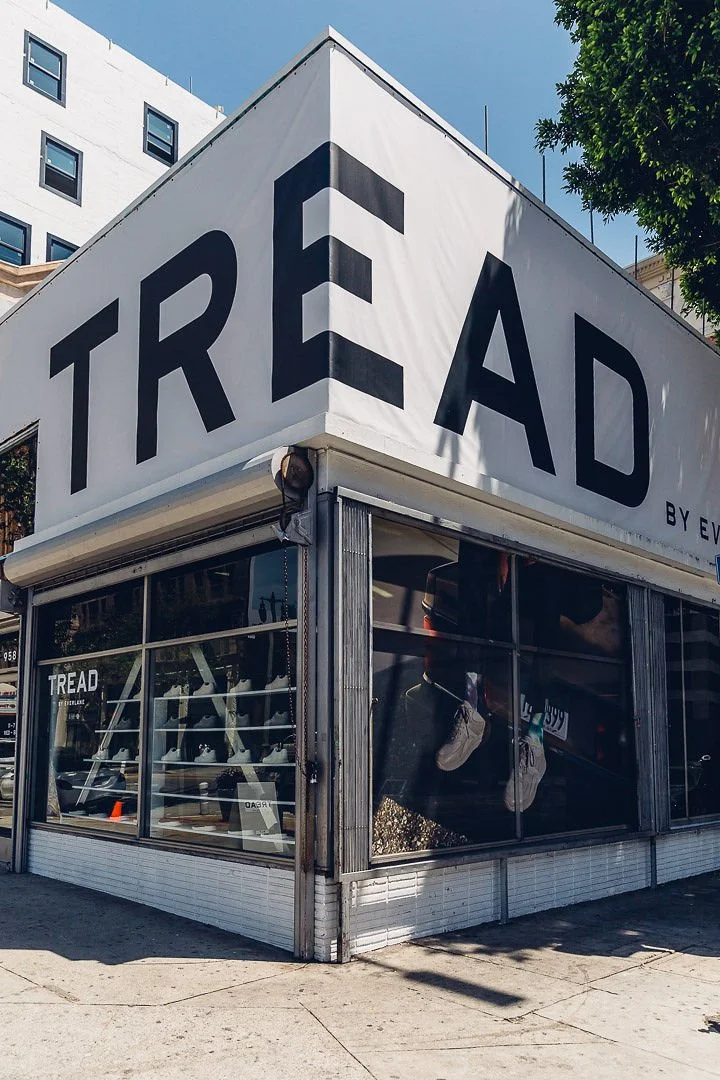 Corner of a retail store with a large sign reading 'TREAD' on the top and a window display with sneakers, with a building and blue sky in the background.