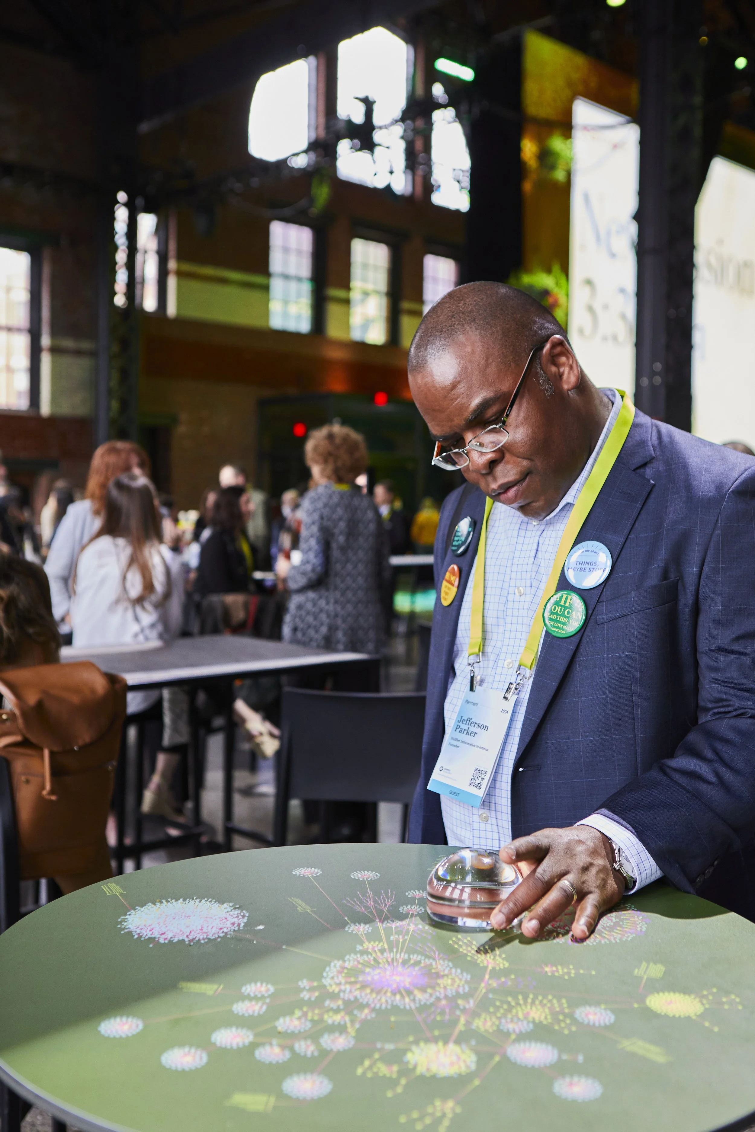 A man in a suit with glasses and conference badges looking down at a circular table with a colorful display of fireworks or starburst graphic. There are several people in the background at an indoor event, with high windows and industrial architecture.