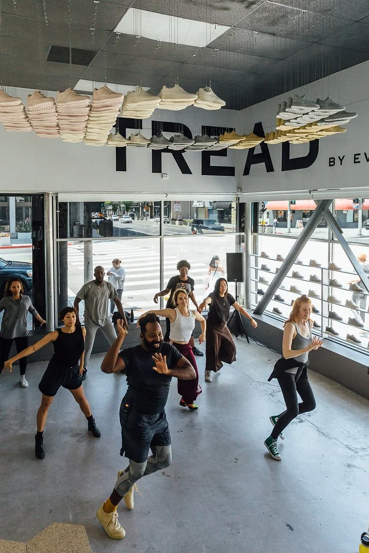 Group of people dancing together inside a shoe store named Tread, with shoes displayed on shelves near large windows and an art installation of shoe insoles hanging from the ceiling.