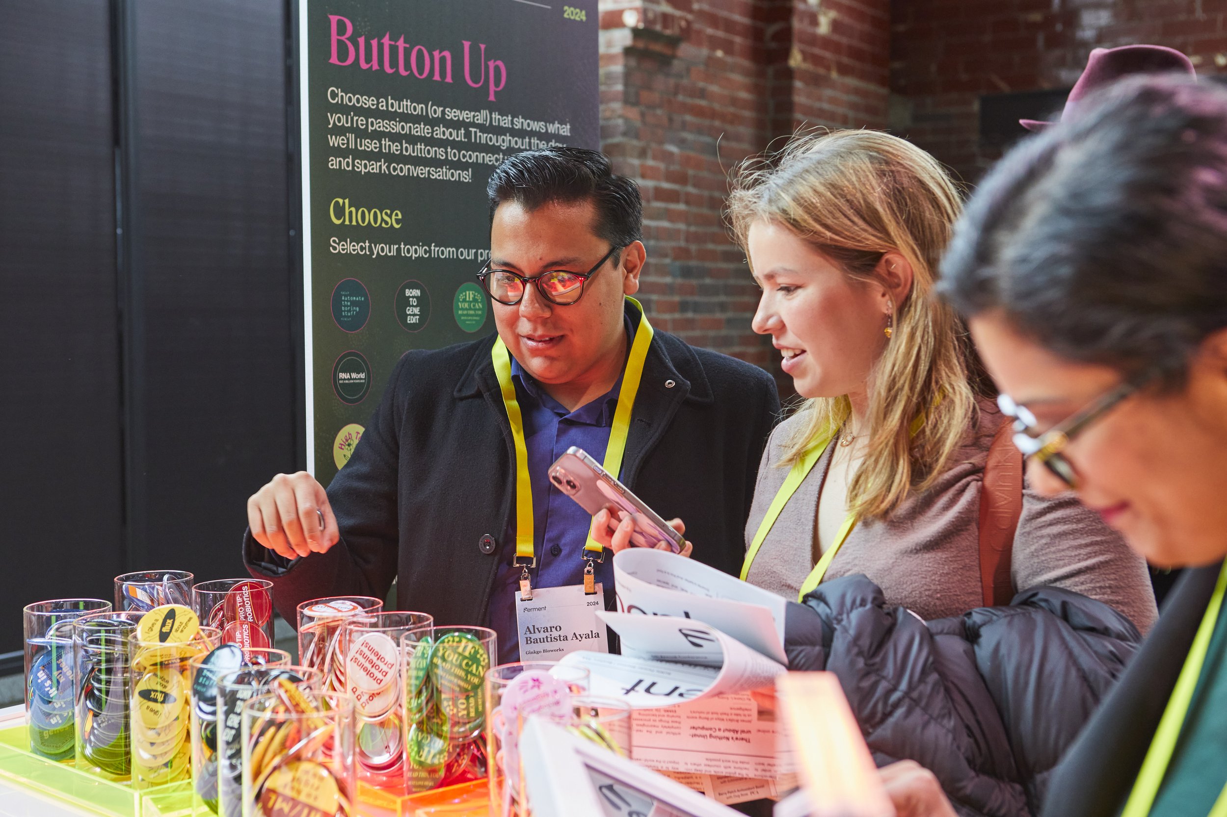 Three people standing behind a table filled with colorful buttons, engaging in conversation and examining items. A poster in the background reads 'Button Up,' with instructions for choosing buttons to connect and spark conversations. The individuals are wearing yellow lanyards and appear to be at an event or exhibition.