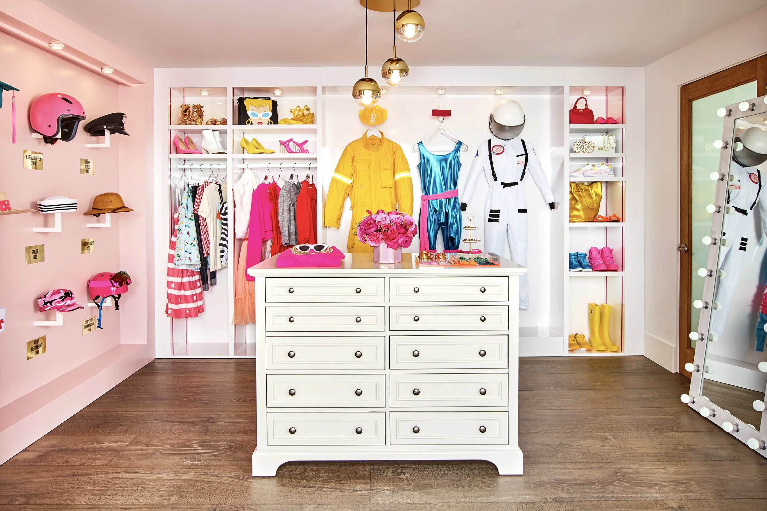 A brightly colored walk-in closet with shelves of clothing, shoes, and accessories, a central white dresser, and a full-length mirror with lights.