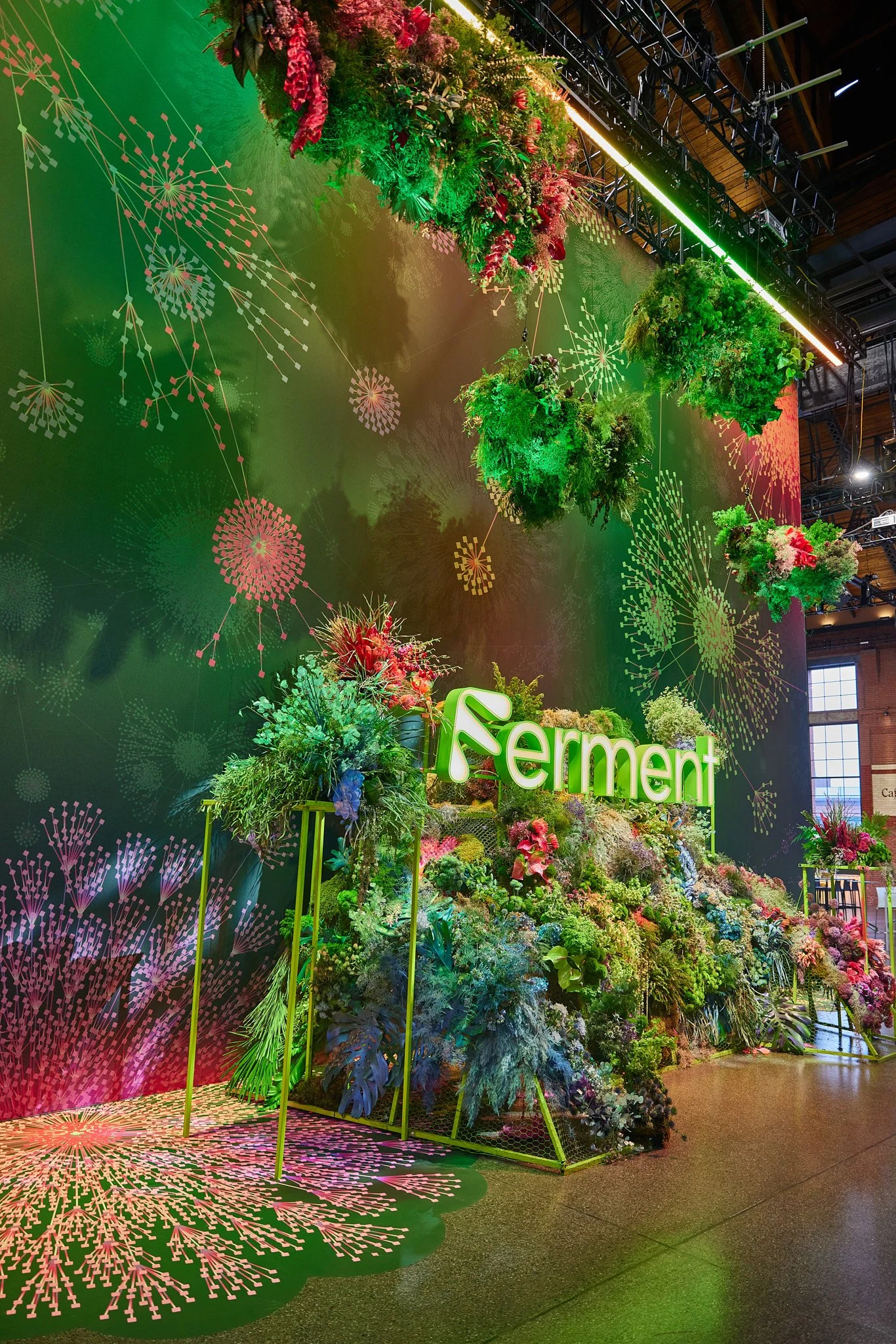 Indoor display with a large green wall decorated with pink visual patterns resembling fireworks, and hanging floral arrangements with various green plants and vibrant flowers. The setup includes the word 'Ferment' in illuminated green letters, with bright lighting accentuating the floral display.