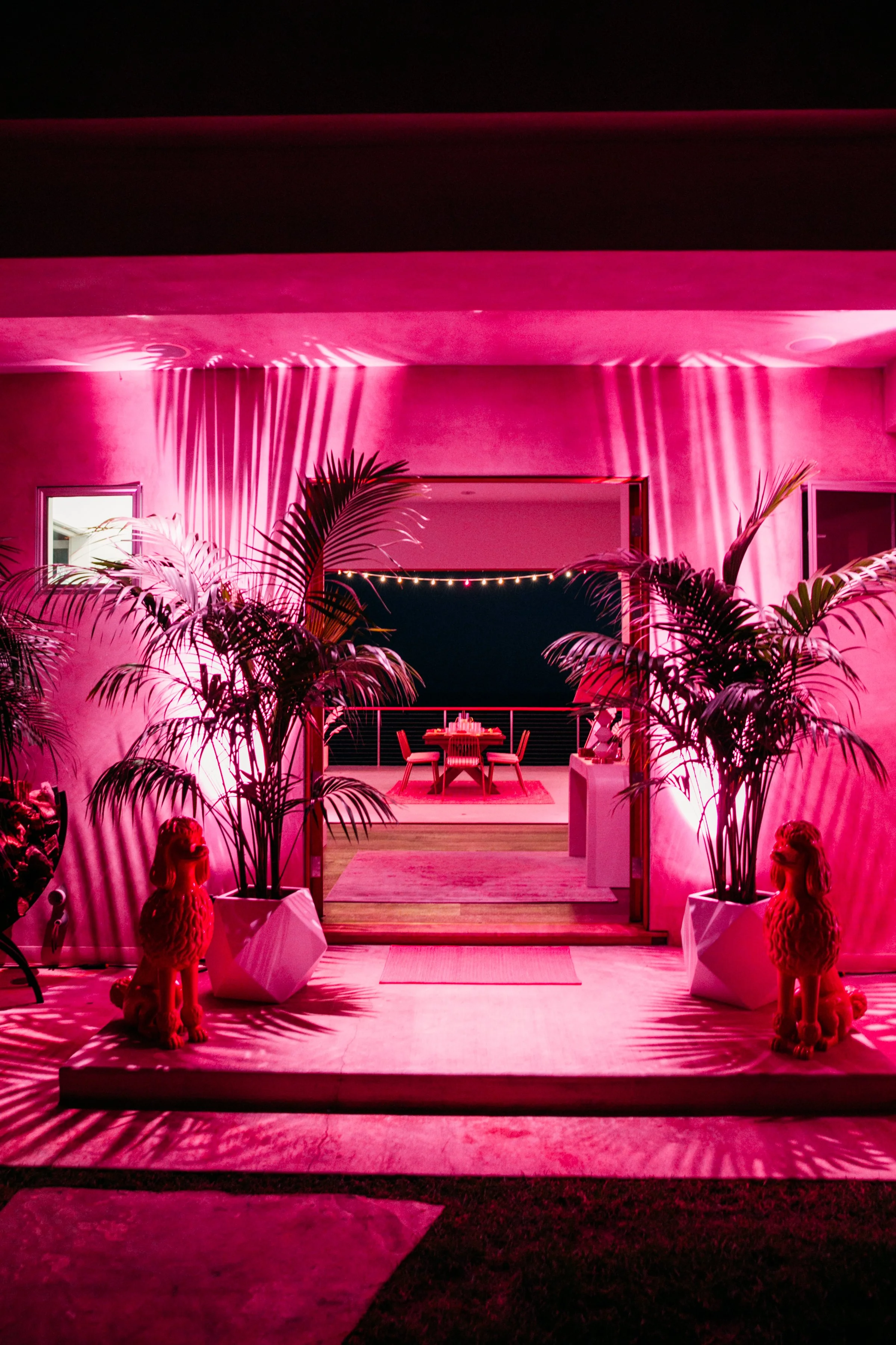 Night scene of a pink-lit outdoor patio area with a dining table and chairs, surrounded by potted palm plants, statues, and string lights, with shadows cast on the wall.